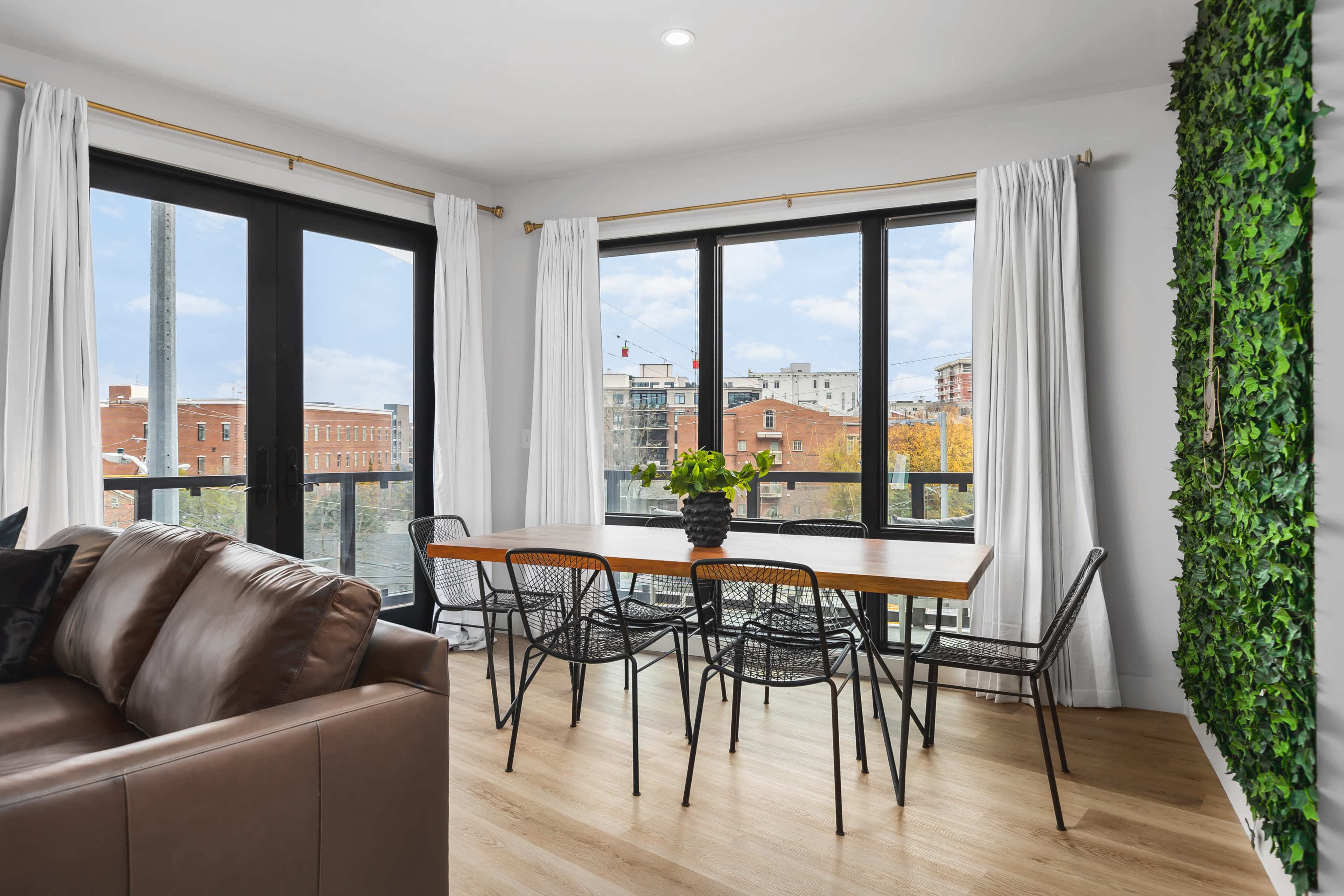 A dining area features a wooden table surrounded by black wire chairs, with large windows offering a view of the city and greenery on one wall.