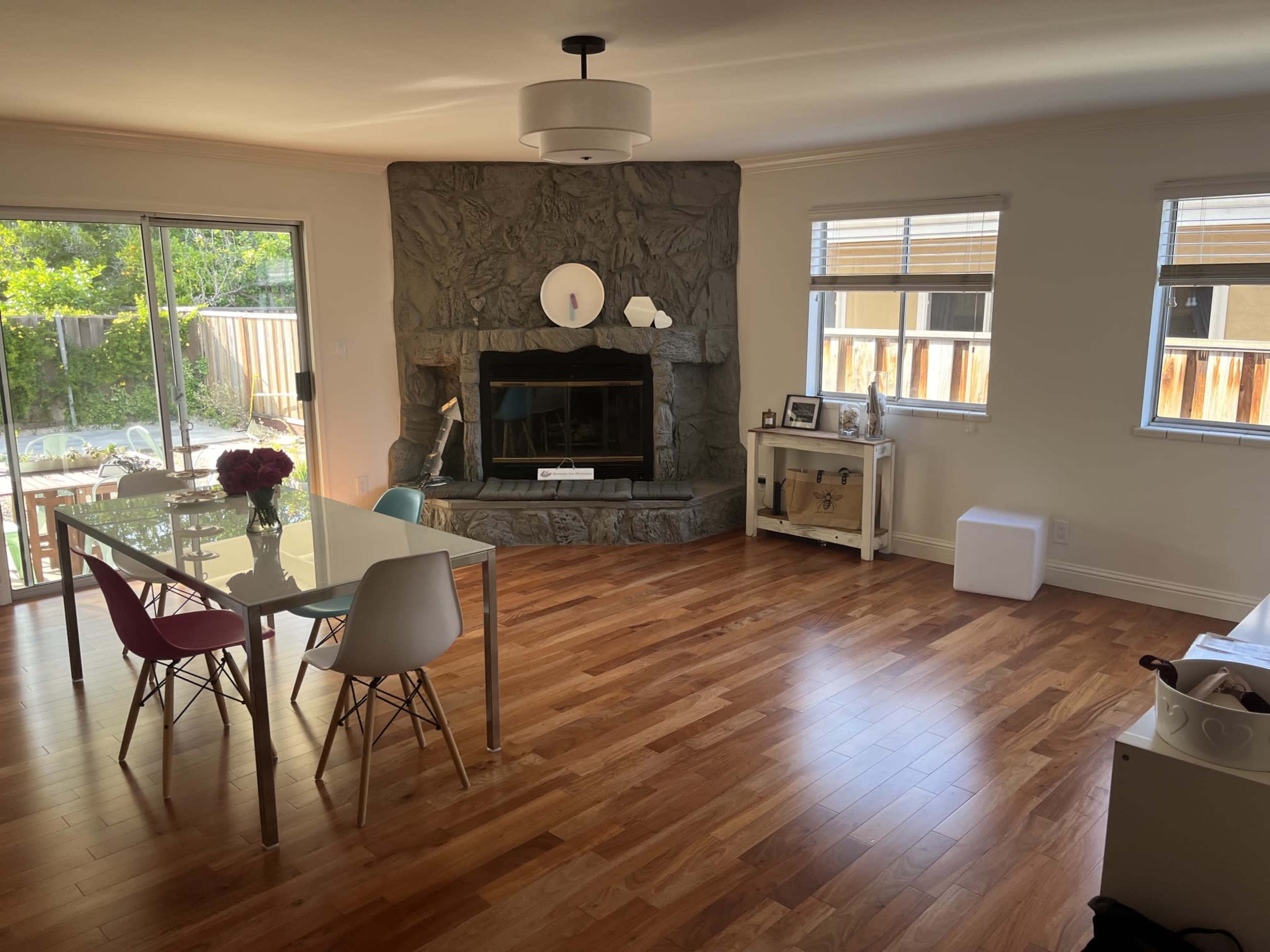The room features a dining area with a glass table and colorful chairs, a stone fireplace, and large windows allowing natural light to enter.