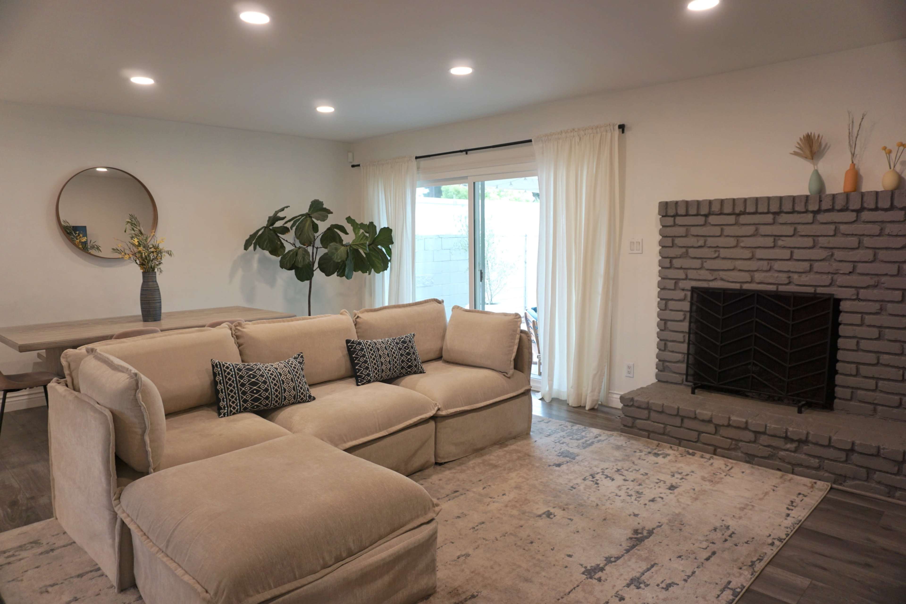 The image shows a modern living room featuring a beige sectional sofa, a round mirror on the wall, a potted plant, and a fireplace made of gray bricks.