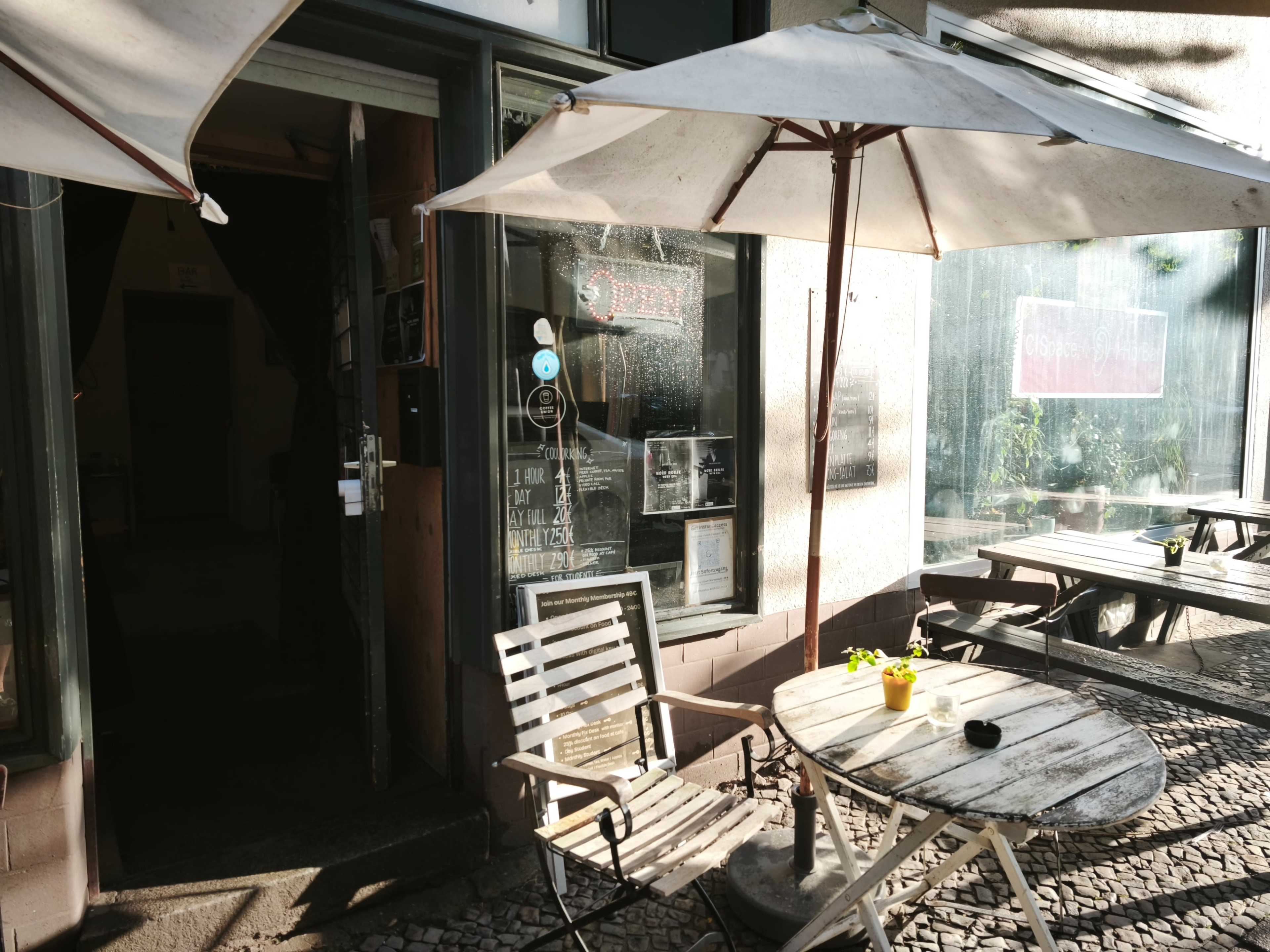 A café with outdoor seating and umbrellas is visible, featuring a wooden table and chairs in front of a glass door.