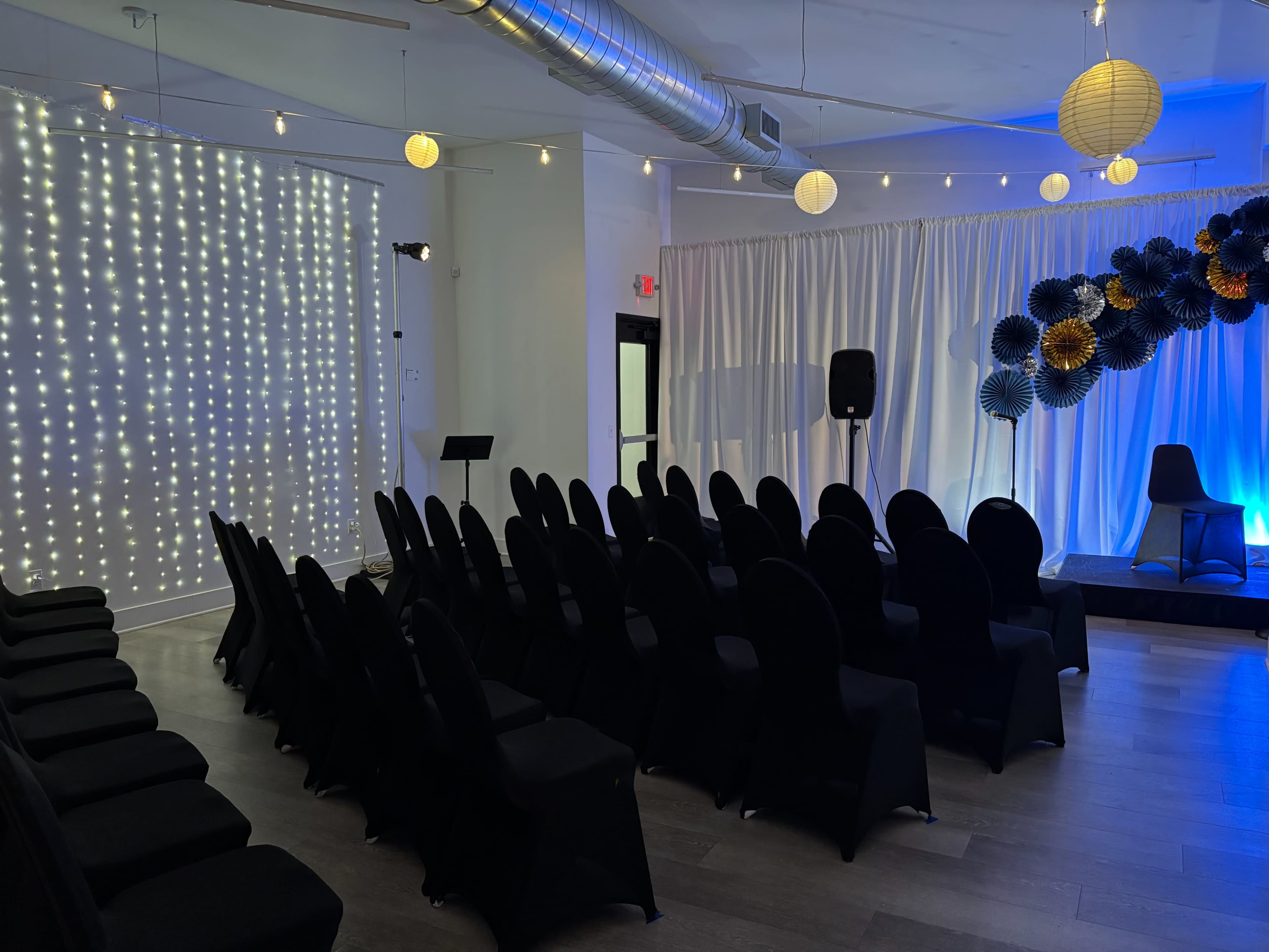The image depicts a room set up for an event, featuring rows of black chairs facing a decorated backdrop with string lights and paper decorations.