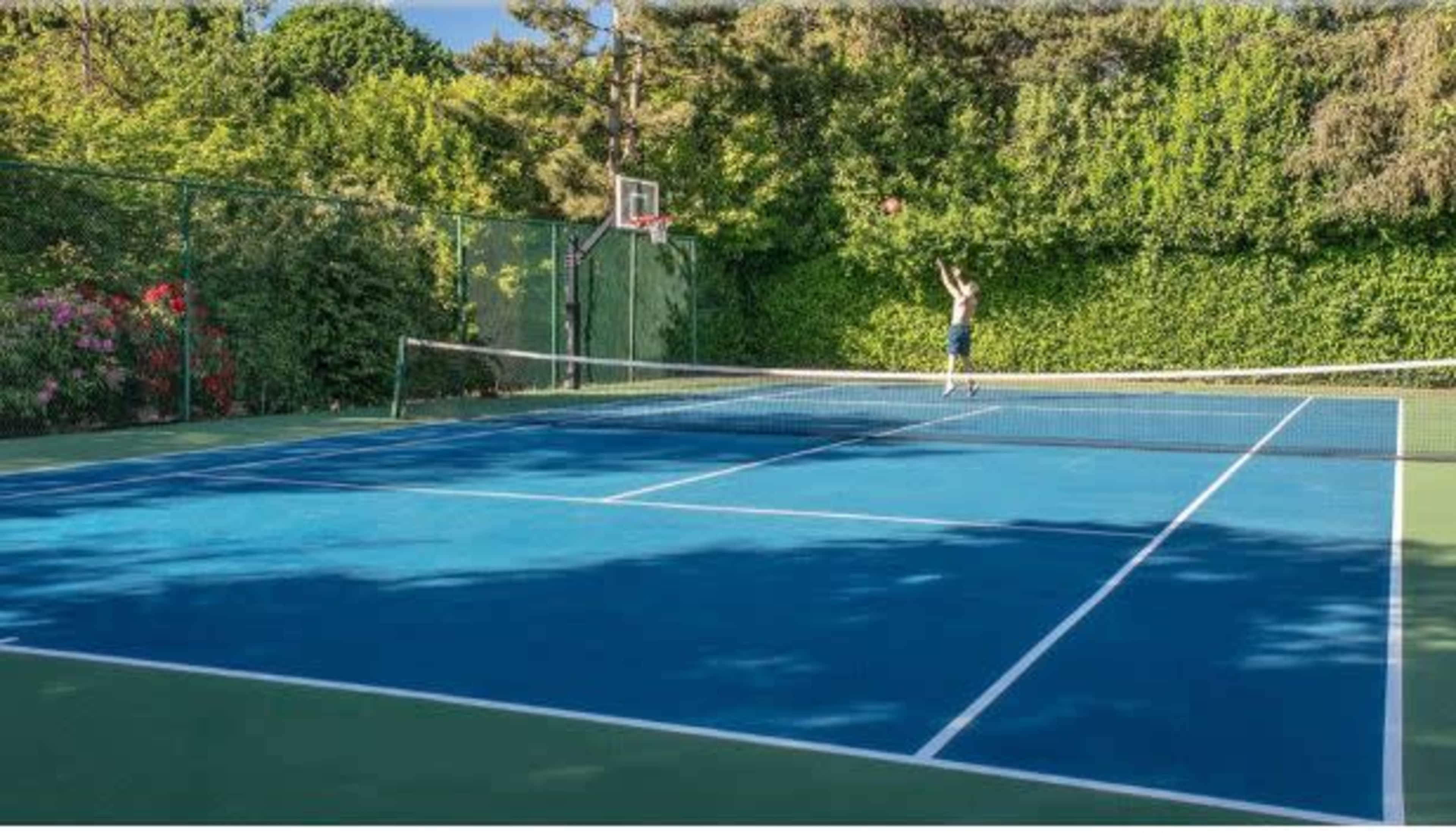A person is shooting a basketball on a court adjacent to a tennis court surrounded by greenery.
