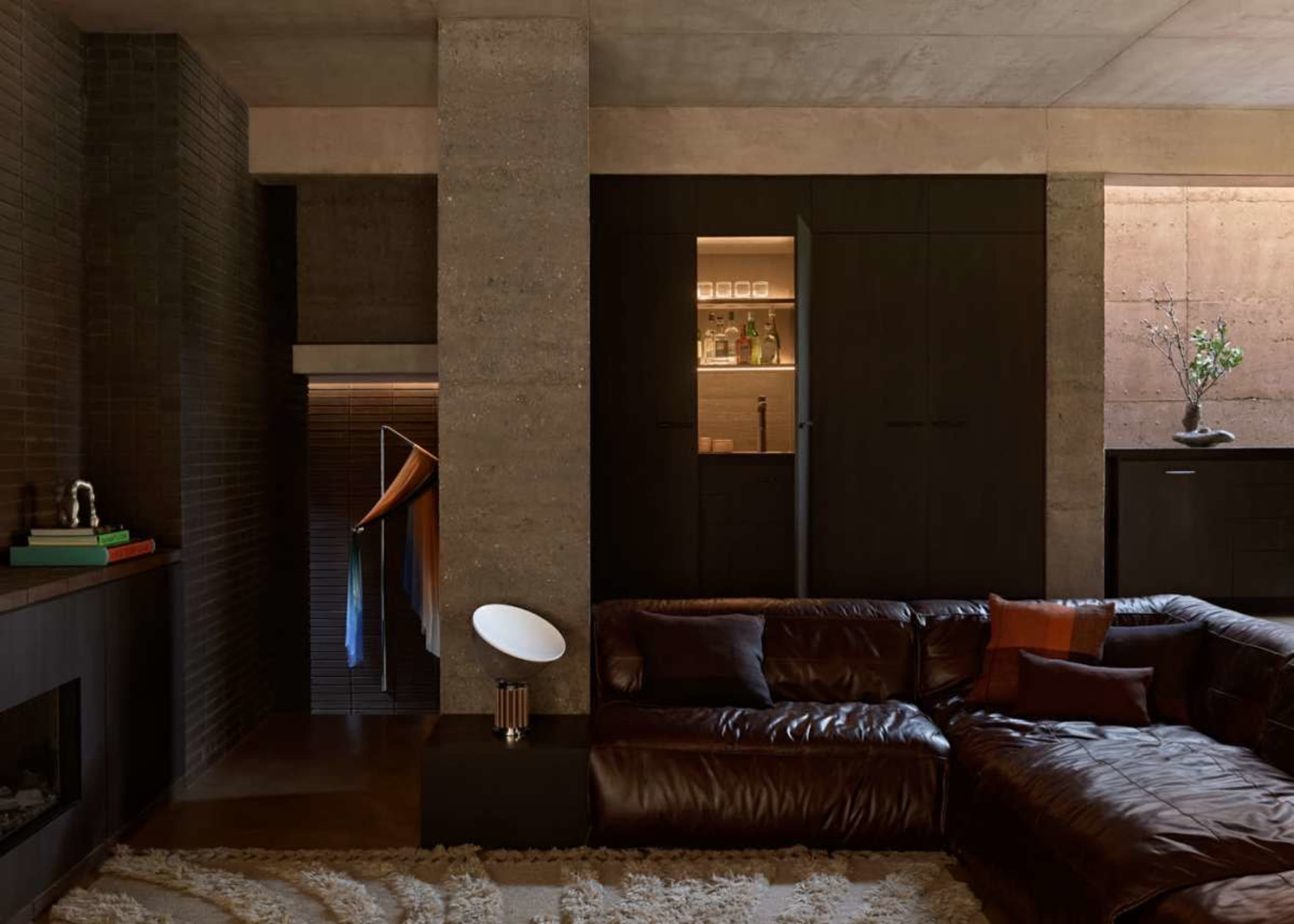 The image showcases a modern living room featuring a large brown leather sectional sofa, a concrete pillar, and a minimalist kitchen area with dark cabinetry.