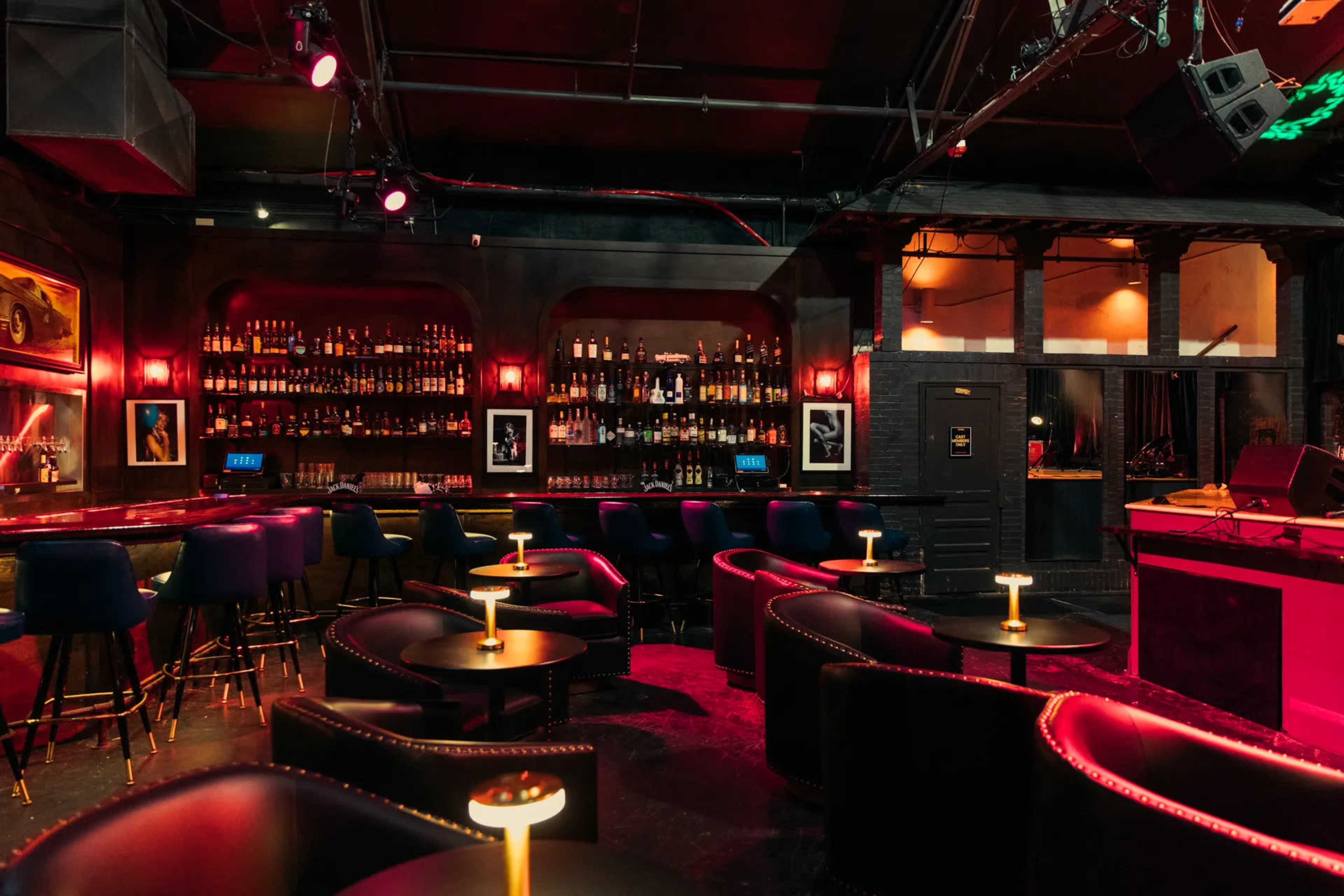 A dimly lit bar features a long shelf of bottles in the background, with small tables and dark chairs arranged in a cozy layout.