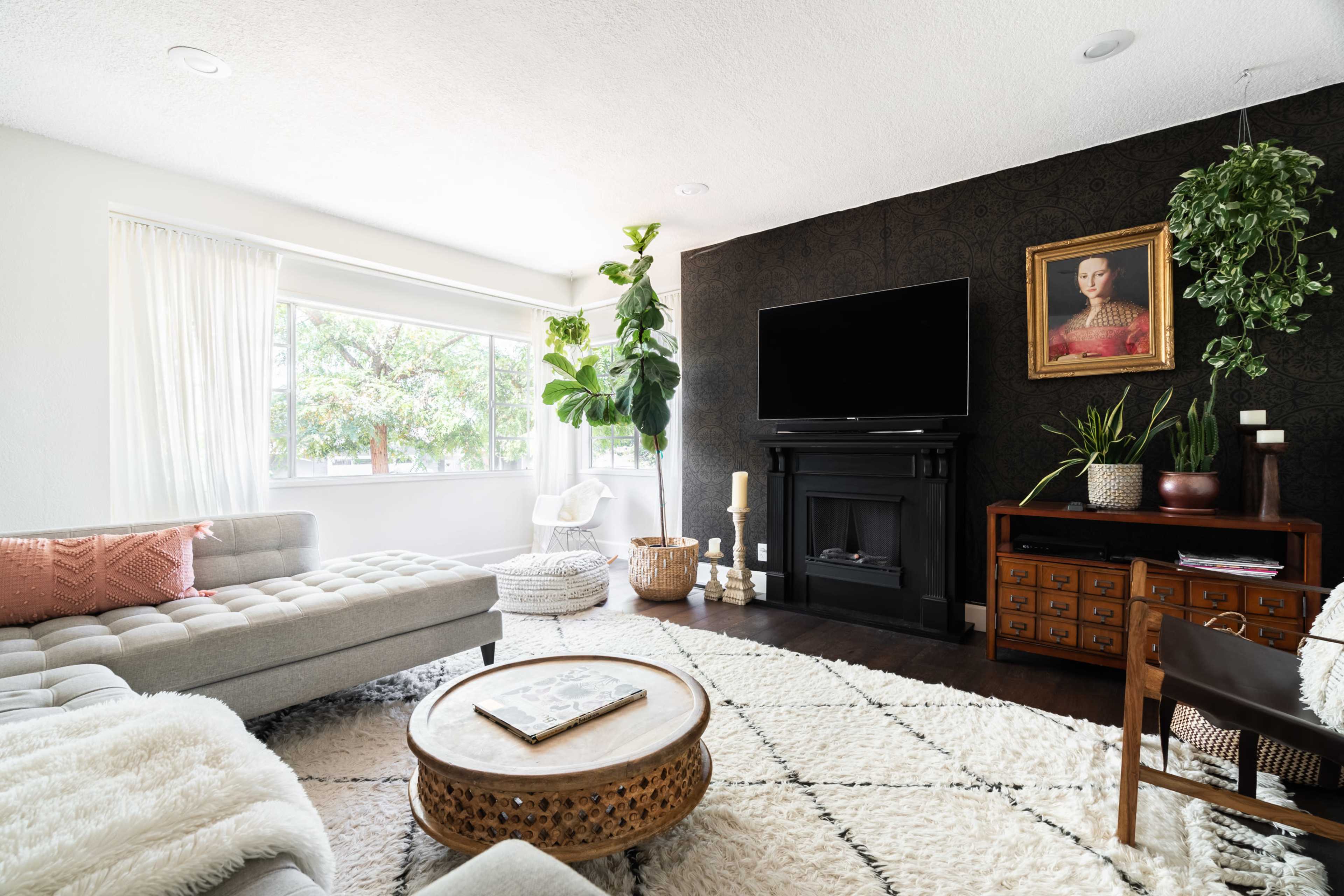 A modern living room features a large sectional sofa, a round coffee table, a flat-screen TV mounted above a fireplace, and vibrant indoor plants.