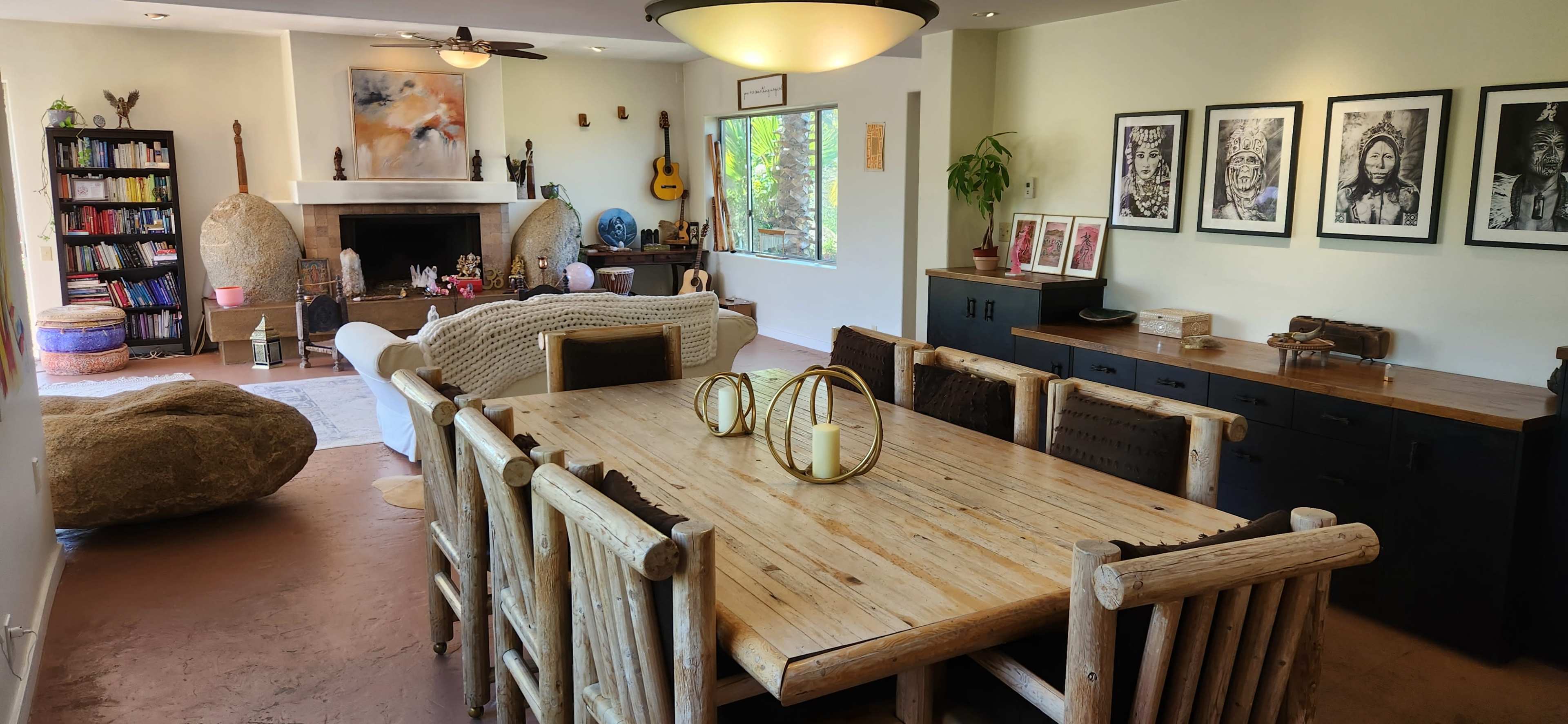A spacious living area with a large wooden dining table, a cozy seating area, and various decorative elements including artwork on the walls and plants by the window.