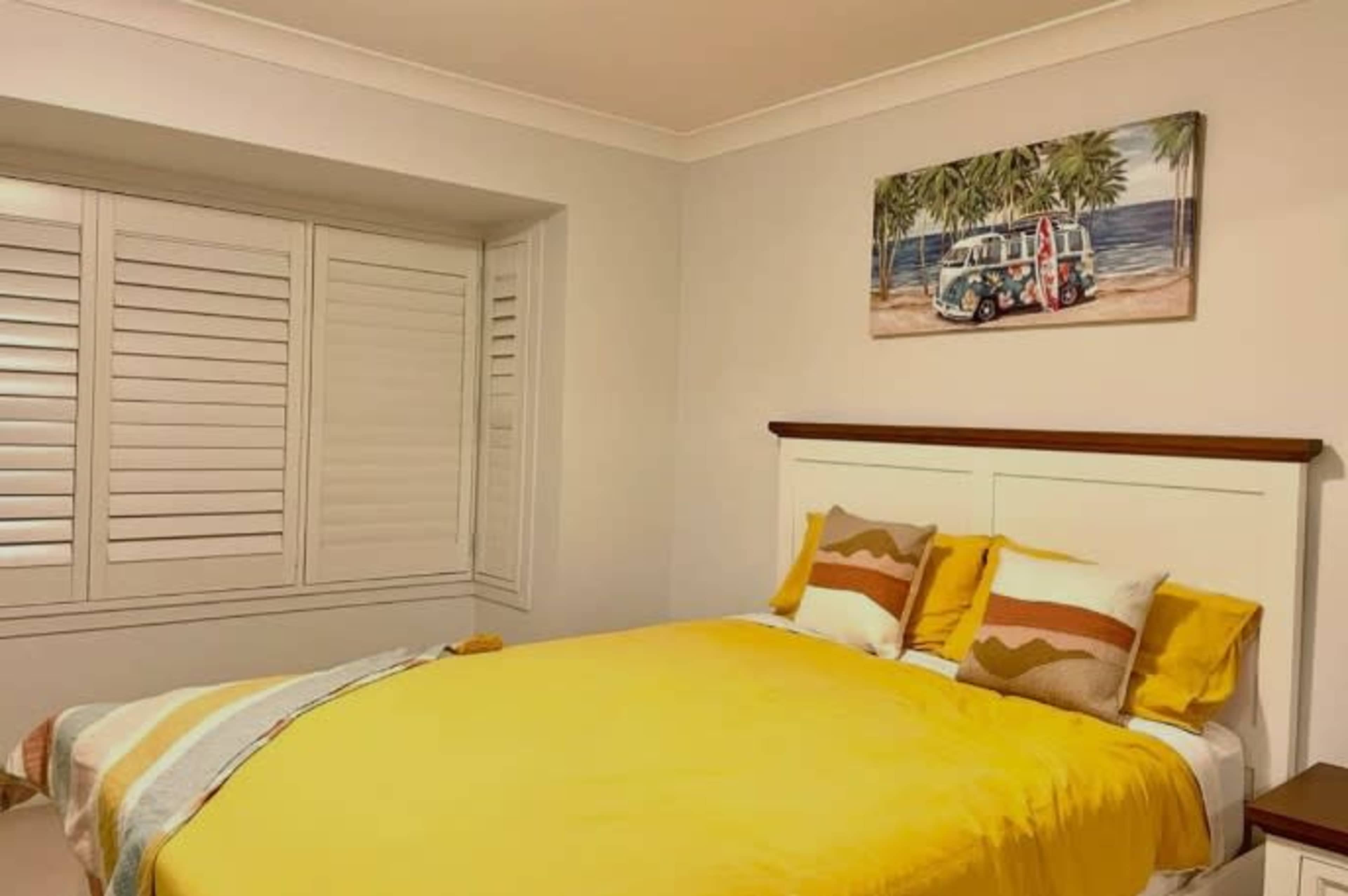 A neatly arranged bedroom with a yellow bedspread and decorative pillows, featuring a painting of a vintage van on the wall above the bed.