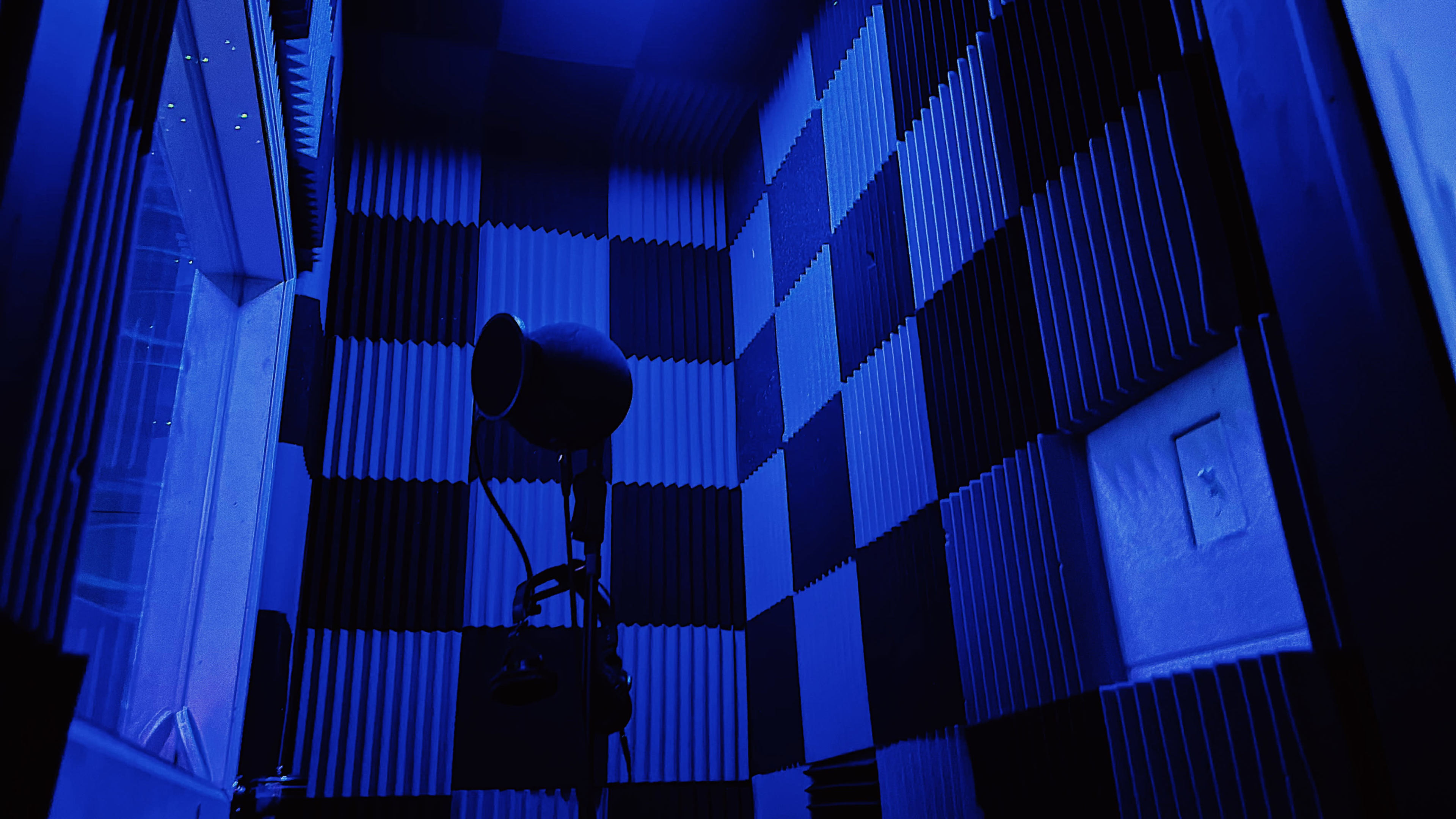 The image shows a soundproof recording booth with blue lighting and acoustic foam panels arranged in a checkered pattern.