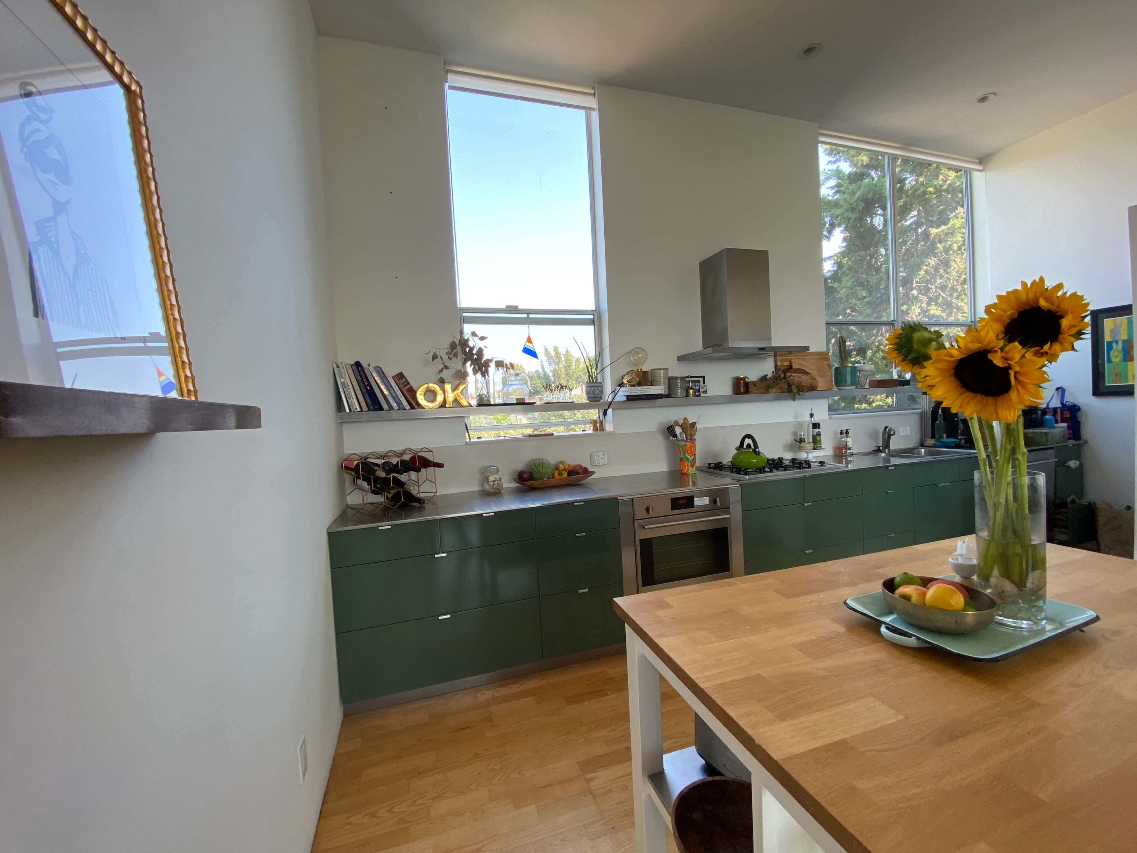 A modern kitchen with green cabinetry, a wooden dining table, and a vase of sunflowers on the table, along with sunlight streaming through large windows.