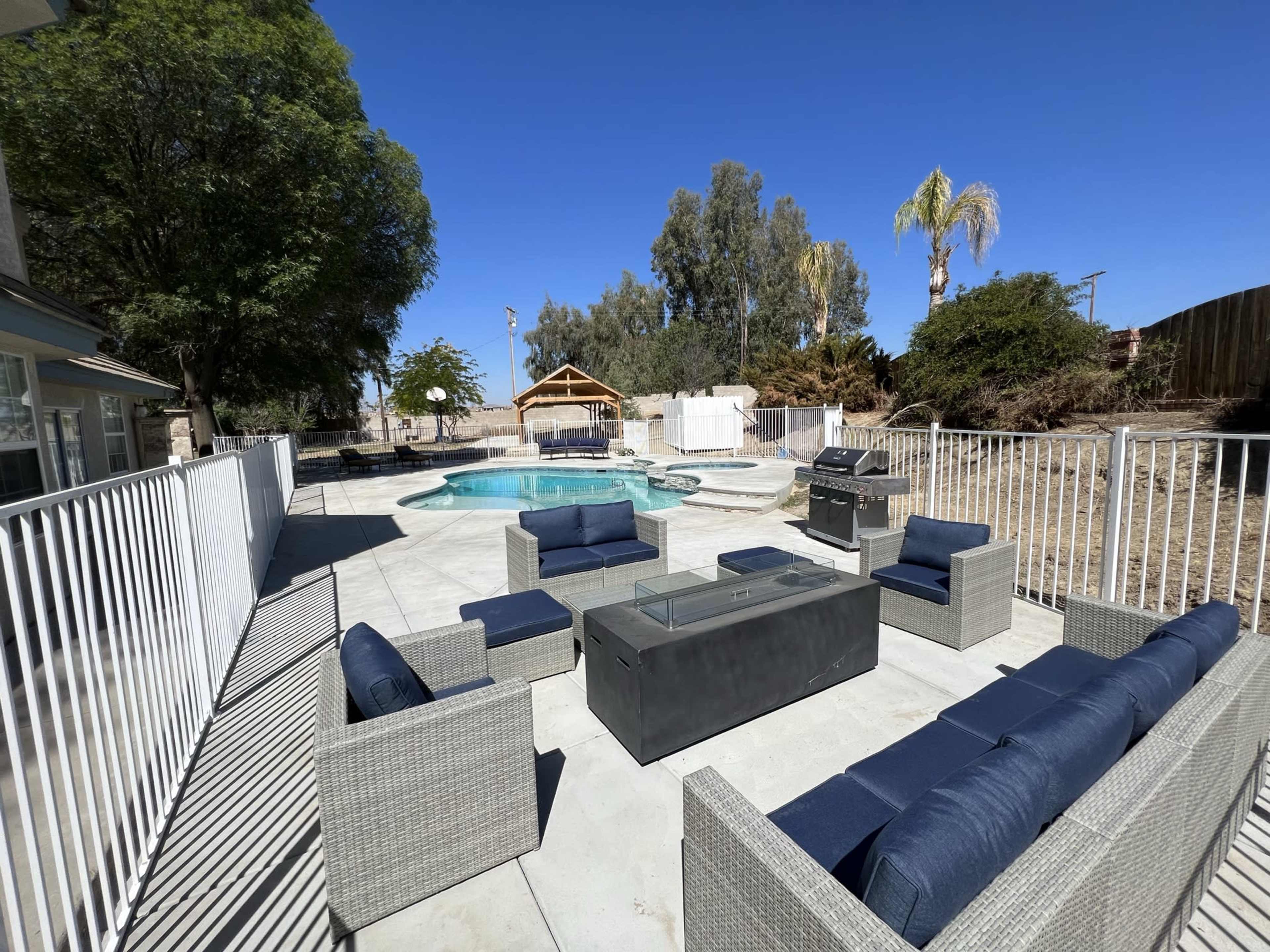 A patio area with outdoor furniture surrounding a pool, enclosed by a white fence and complemented by a nearby gazebo and trees.
