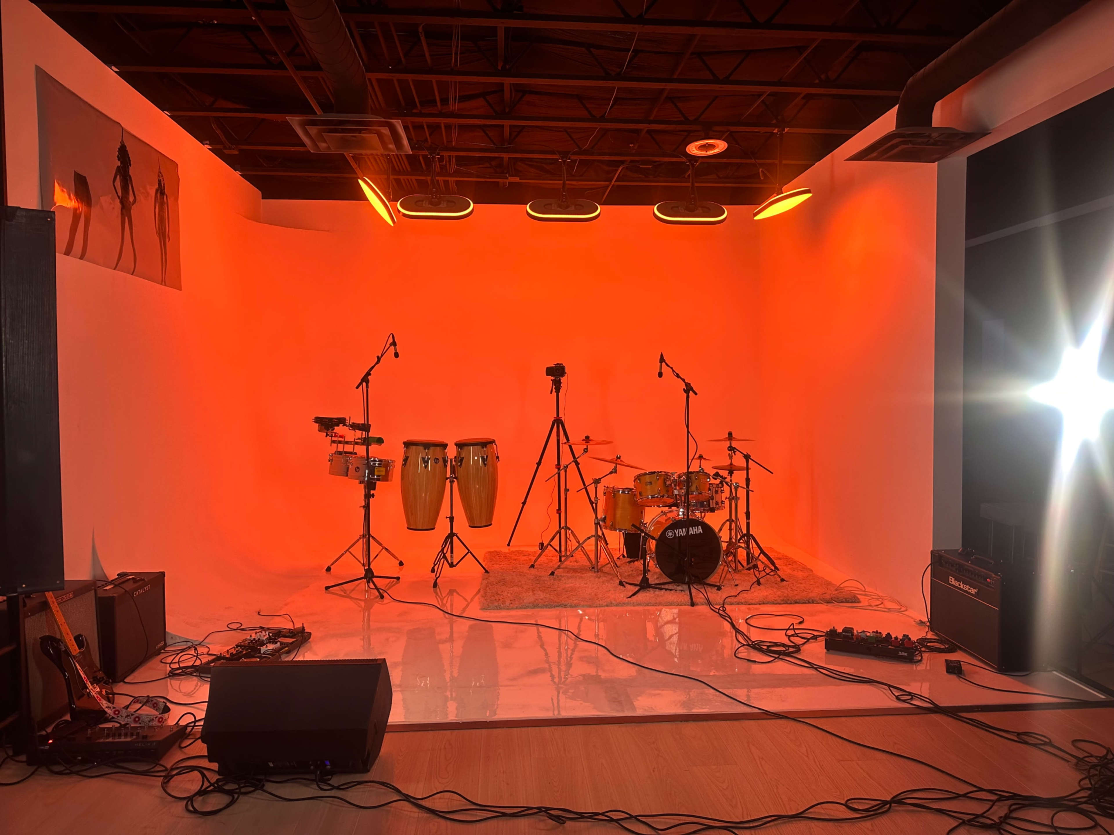 The image shows a music studio setup with various instruments, including congas, a drum set, and microphones, against an orange backdrop.