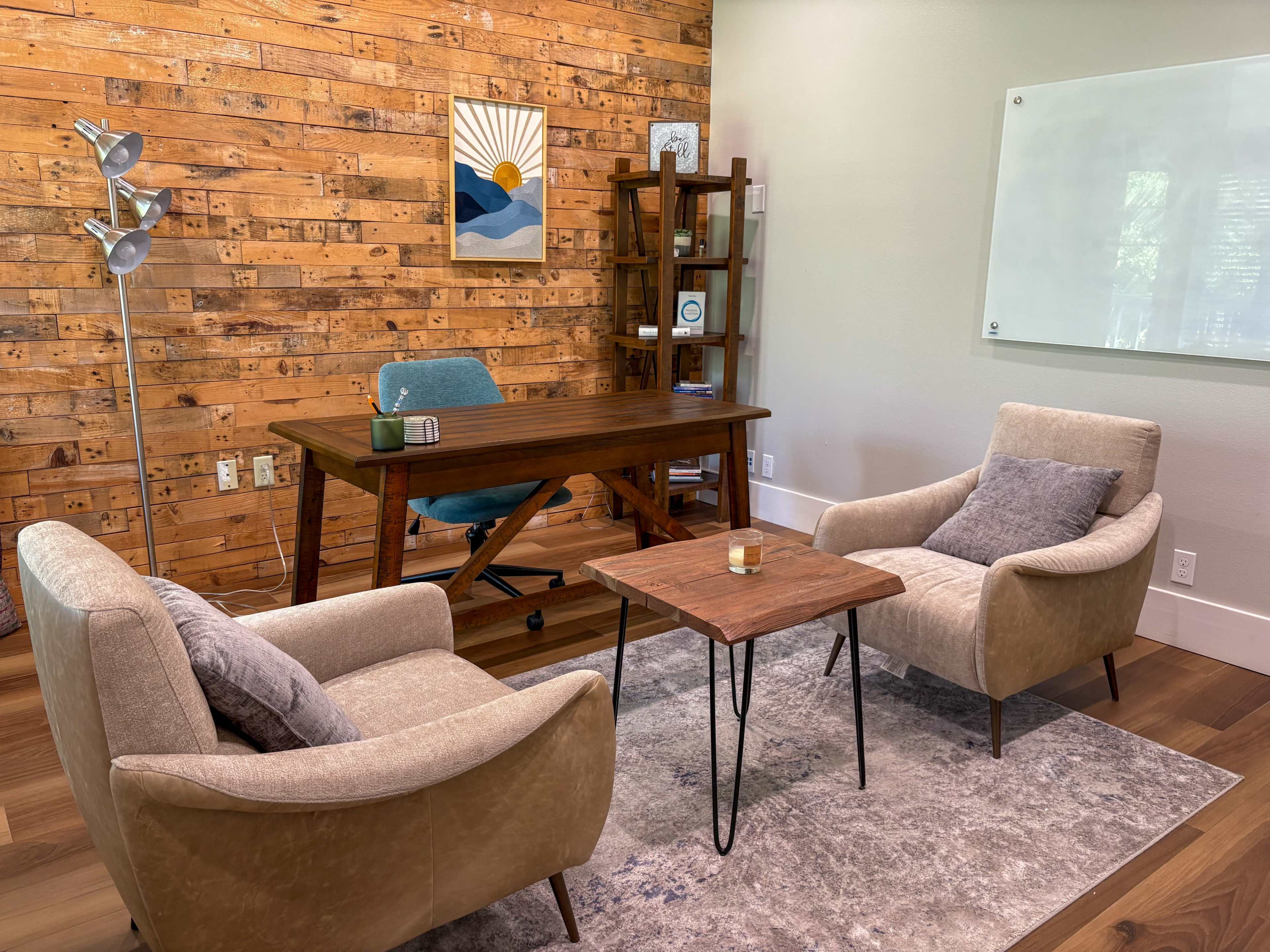 A cozy office space features a wooden desk, two armchairs, a small table, and a wall adorned with wooden panels and artwork.
