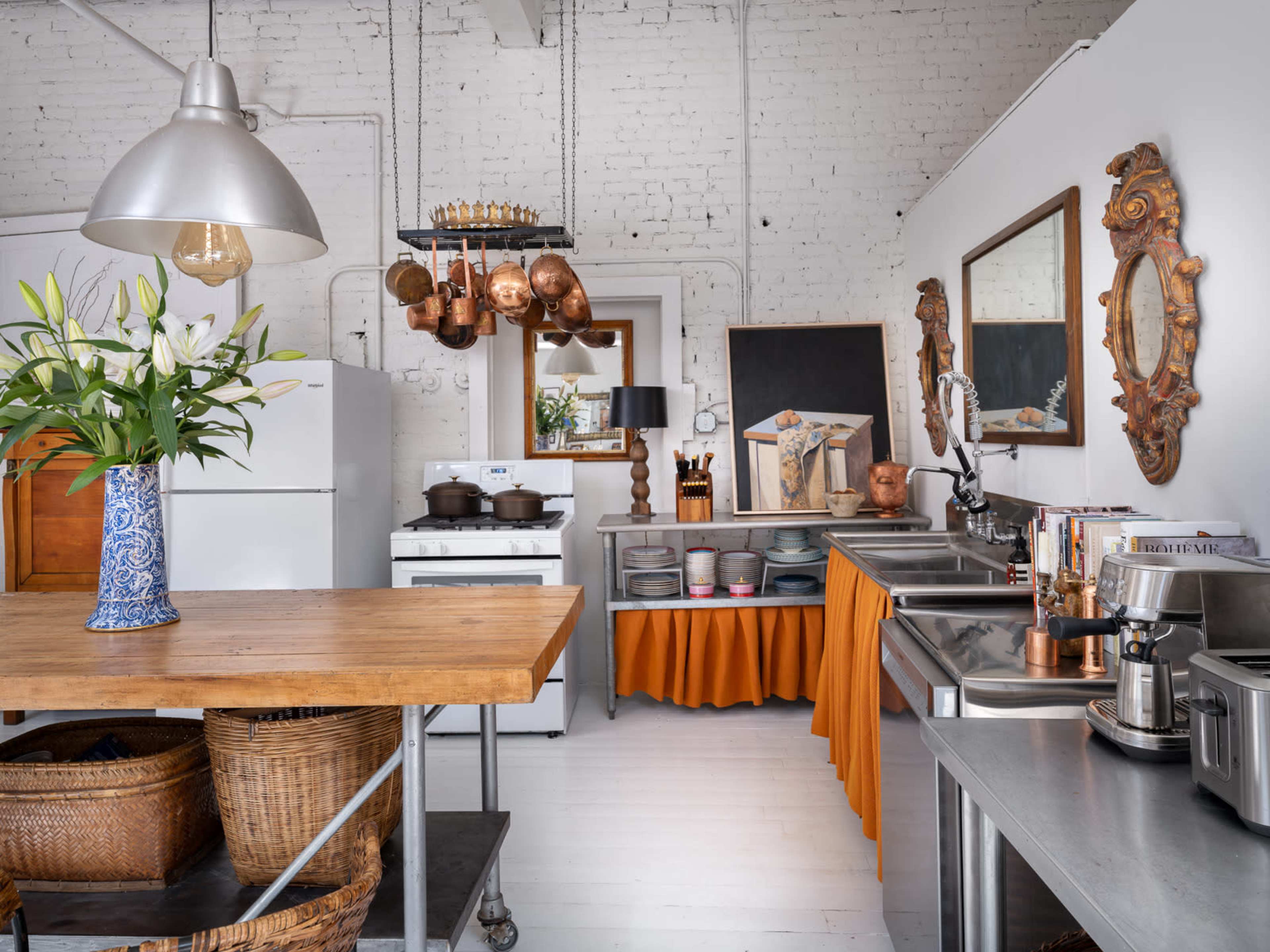 A bright kitchen featuring a central wooden table, stainless steel countertops, a variety of cooking utensils, and decorative elements on the walls.