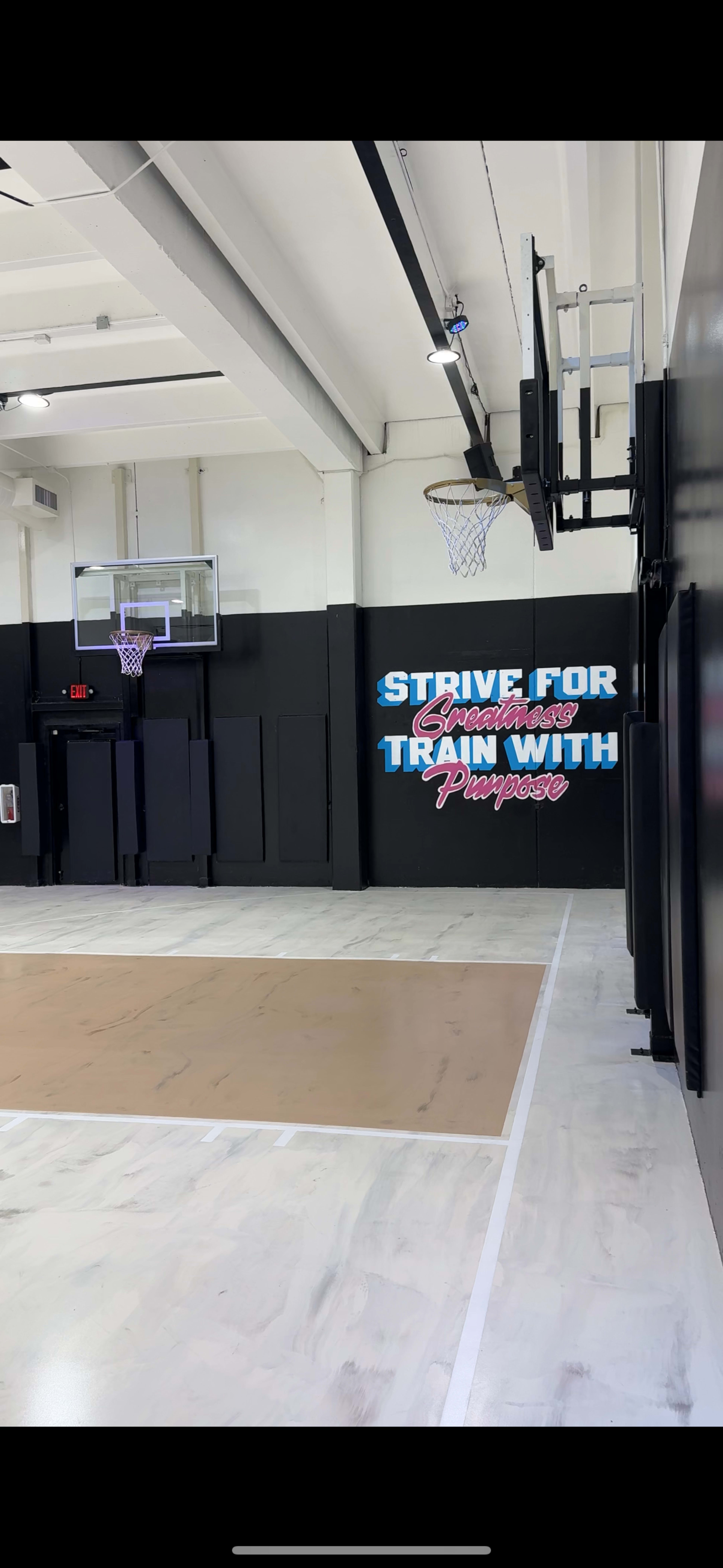 The image shows an indoor basketball court with a hoop on the left and a wall featuring motivational text on the right.