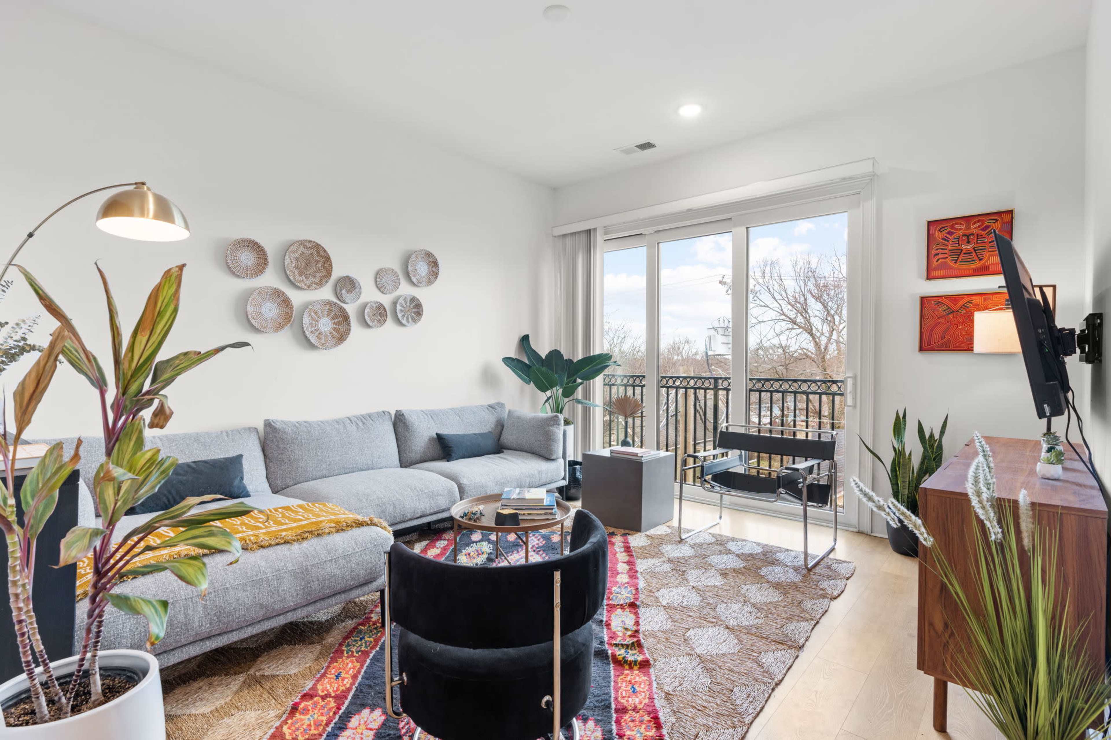 A modern living room features a gray sectional sofa, a round coffee table, and a TV against a white wall with various decorative pieces, complemented by large windows that open to a balcony.