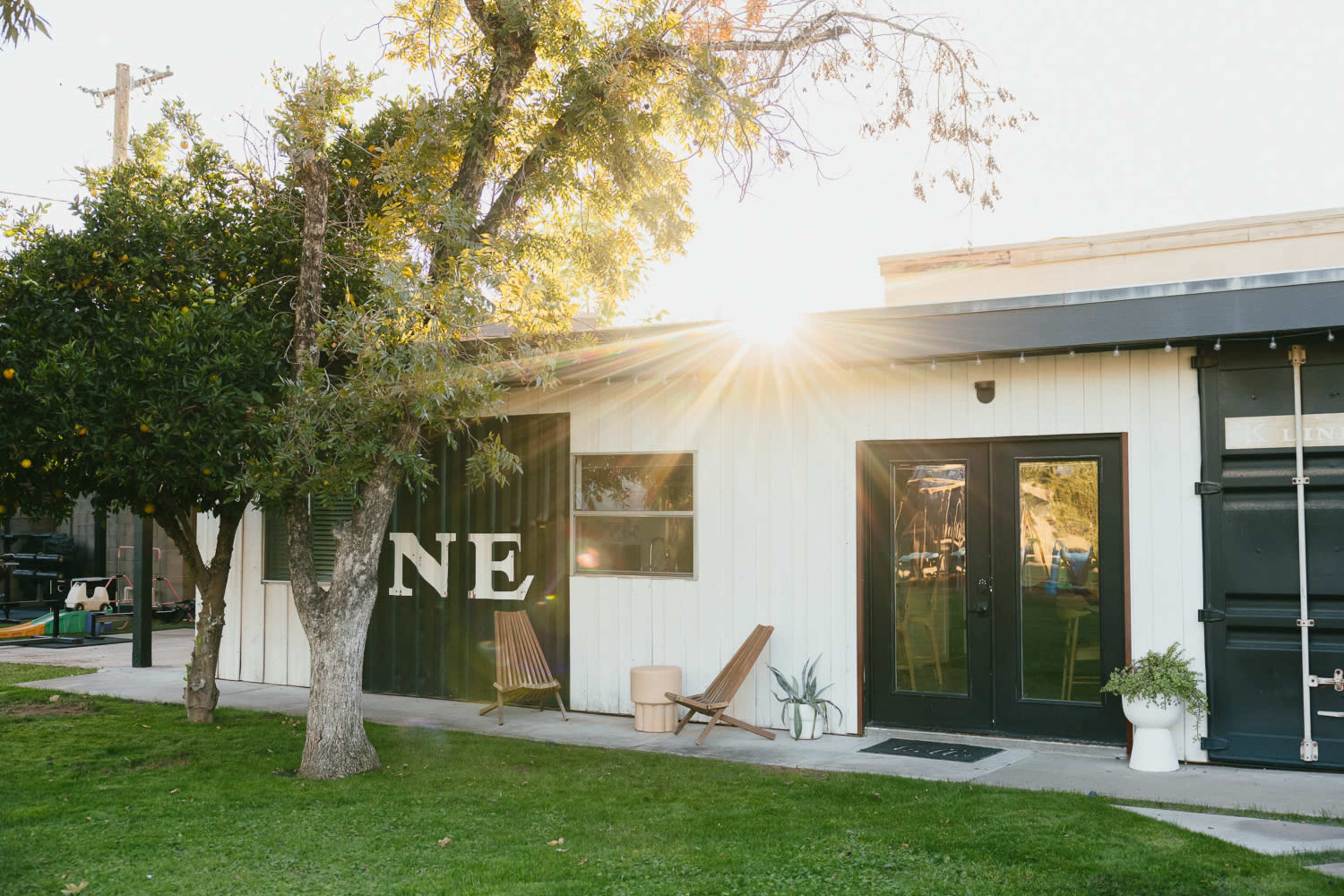 A white building with large black doors and two wooden chairs positioned on a grassy area, with sunlight streaming through trees in the background.