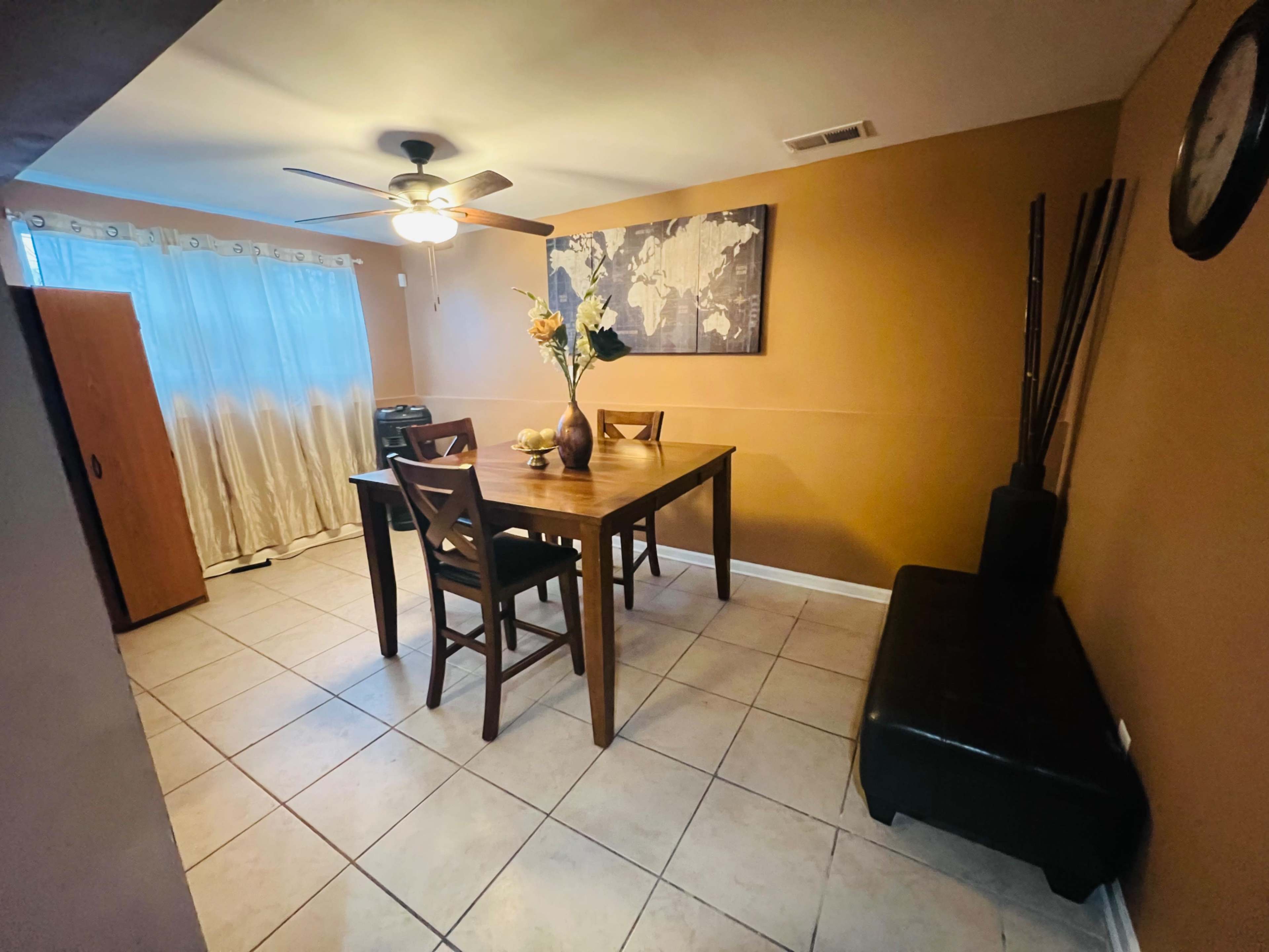 A dining area features a wooden table with chairs, a vase of flowers, a world map on the wall, and a partially visible curtain window.