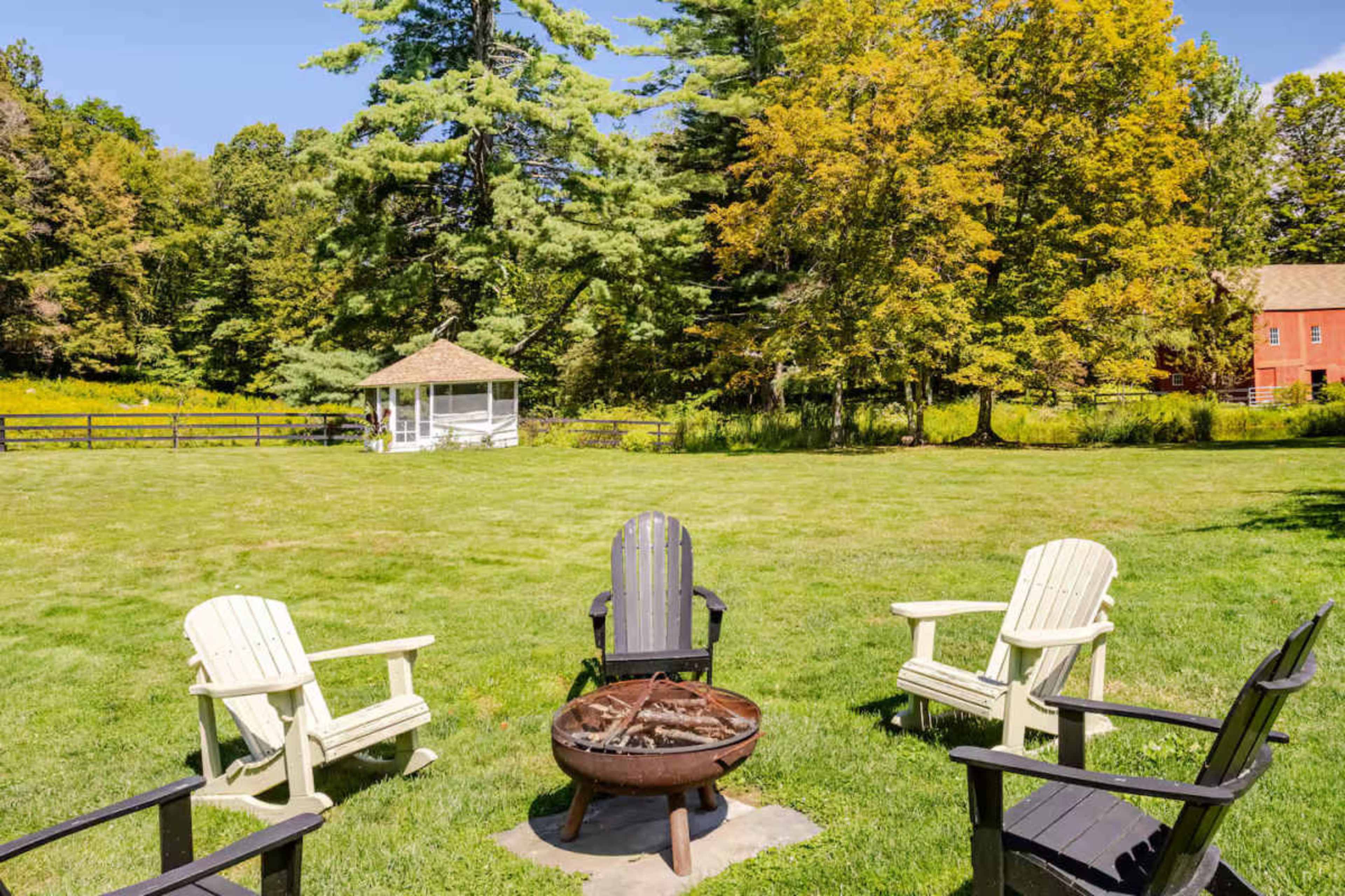 A circular fire pit surrounded by four chairs is positioned on a grassy area with a gazebo and trees in the background.