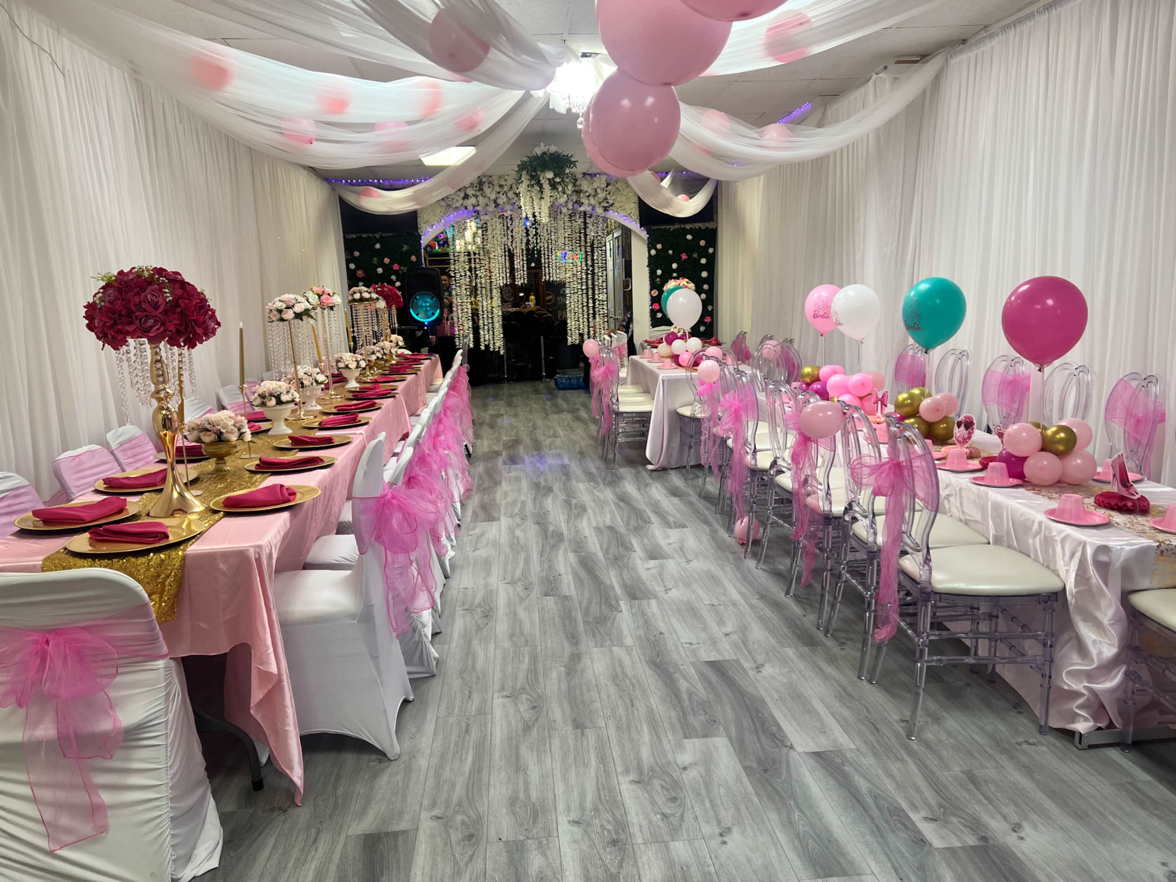 The image shows a decorated event space with multiple tables covered in pink and gold tablecloths, surrounded by balloons and floral arrangements.