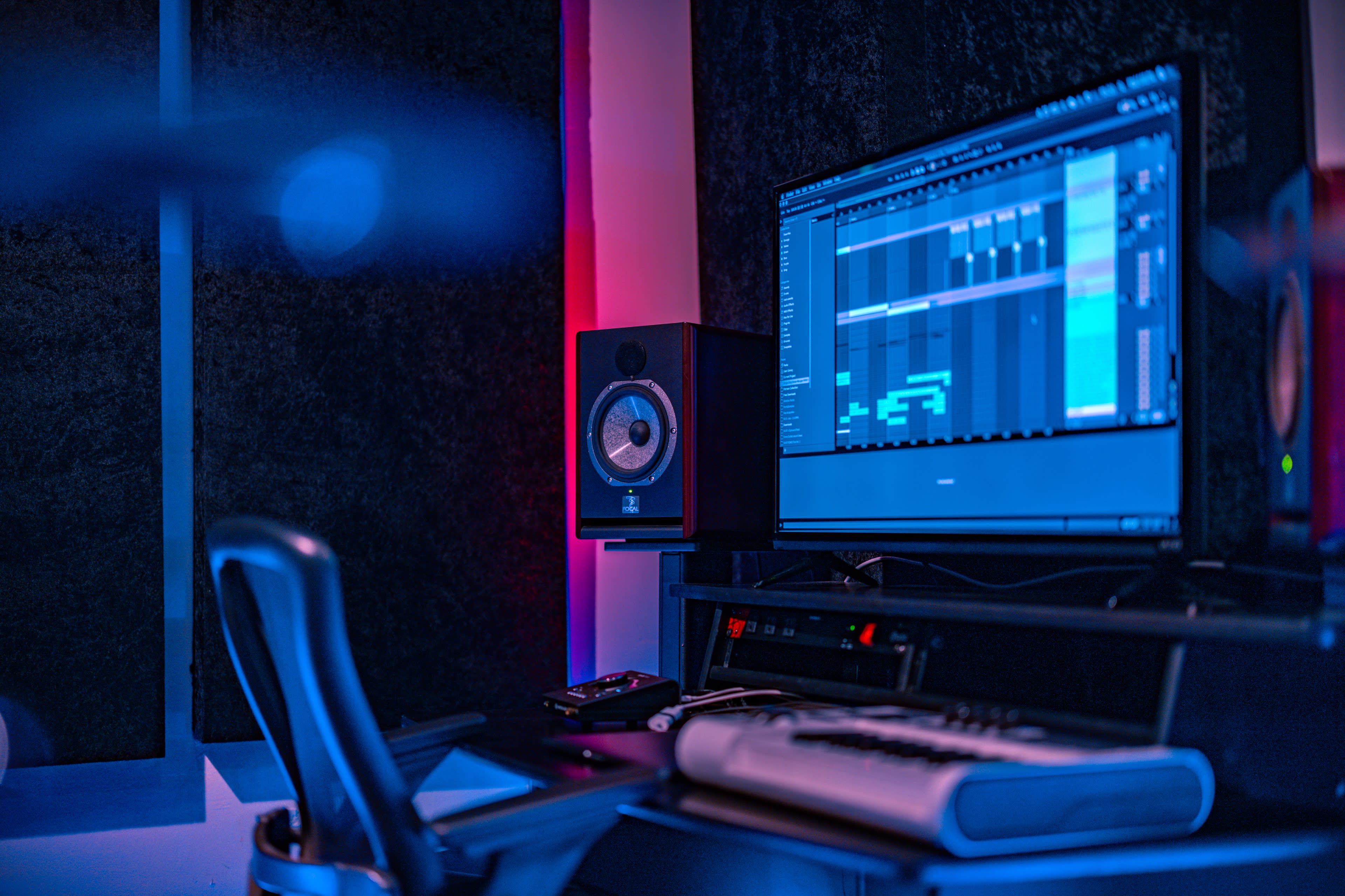A music production setup featuring a computer monitor displaying audio software, a studio monitor speaker, and a MIDI keyboard on a black desk in a soundproofed room.