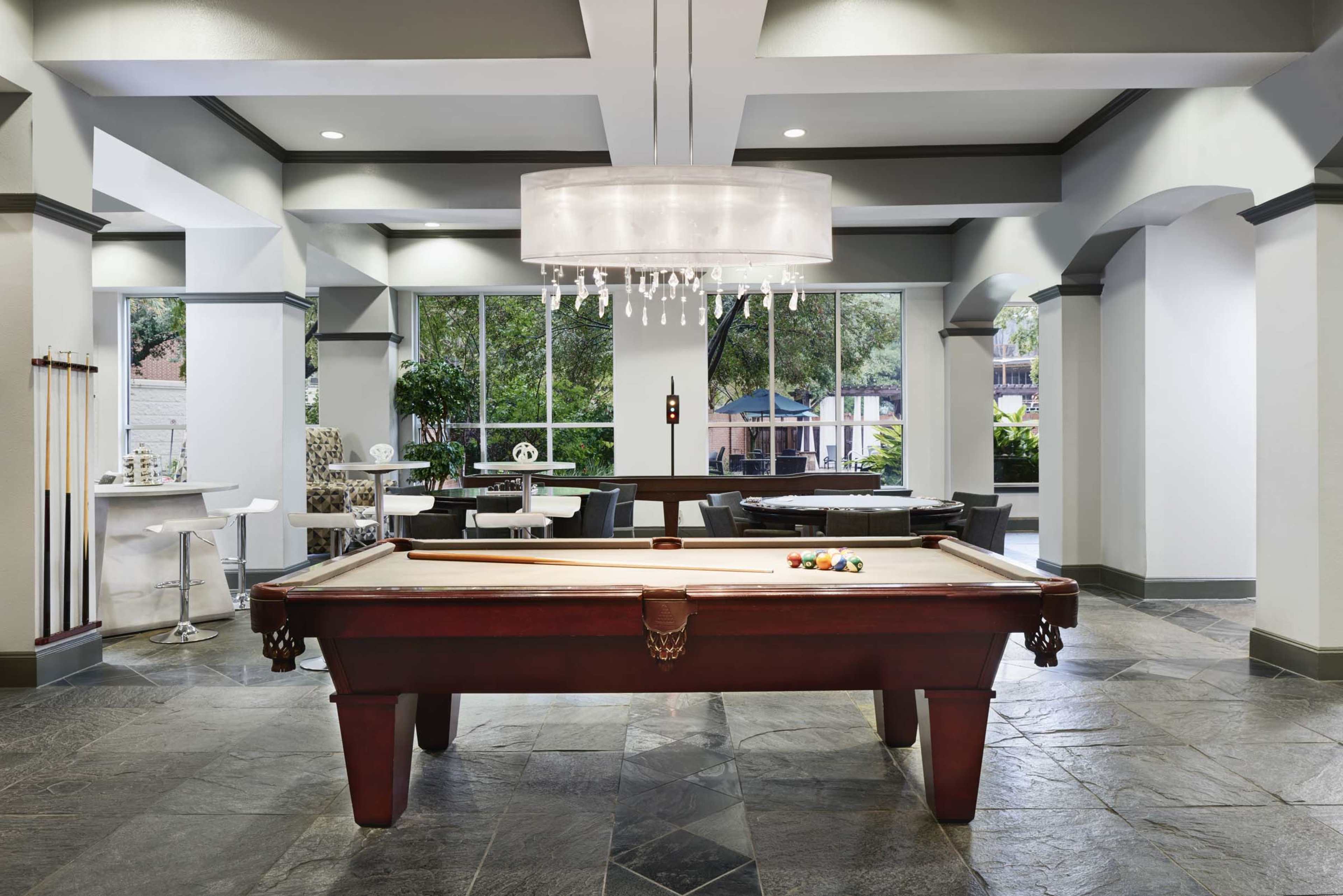 A modern game room featuring a billiard table in the center, surrounded by stylish seating and large windows that let in natural light.