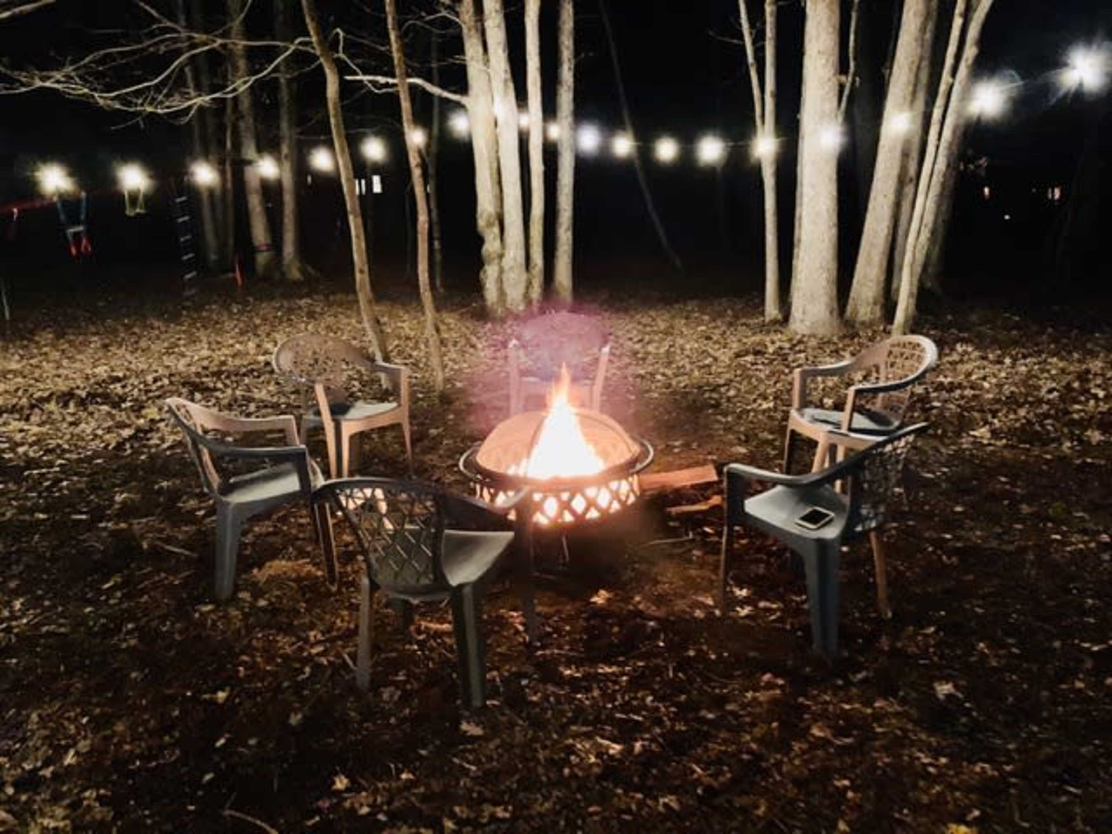 A fire pit surrounded by five chairs sits in a wooded area at night, illuminated by string lights above.