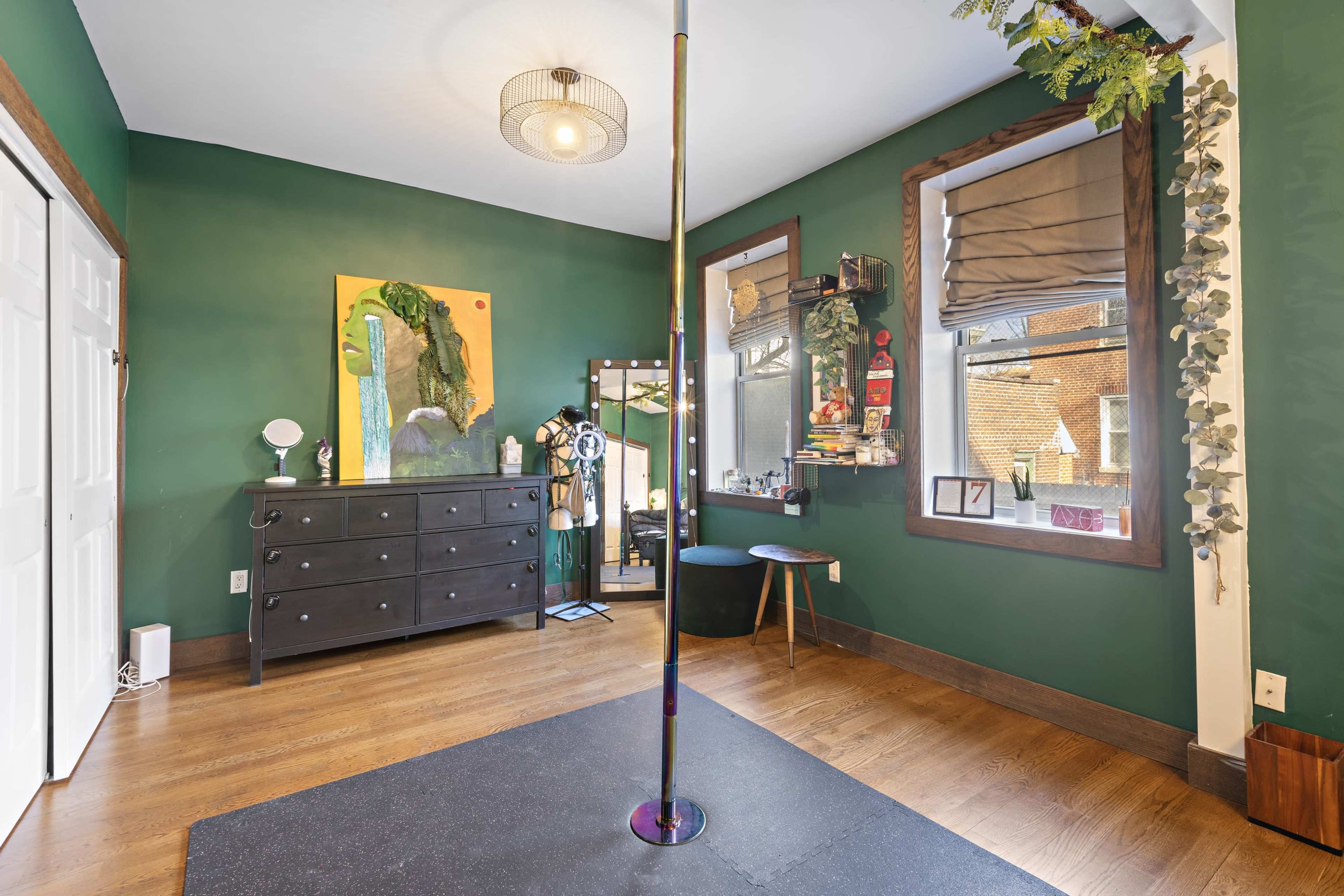 The image shows a room with dark green walls, a dancing pole in the center, a black dresser, a mirror, and two windows adorned with natural light.