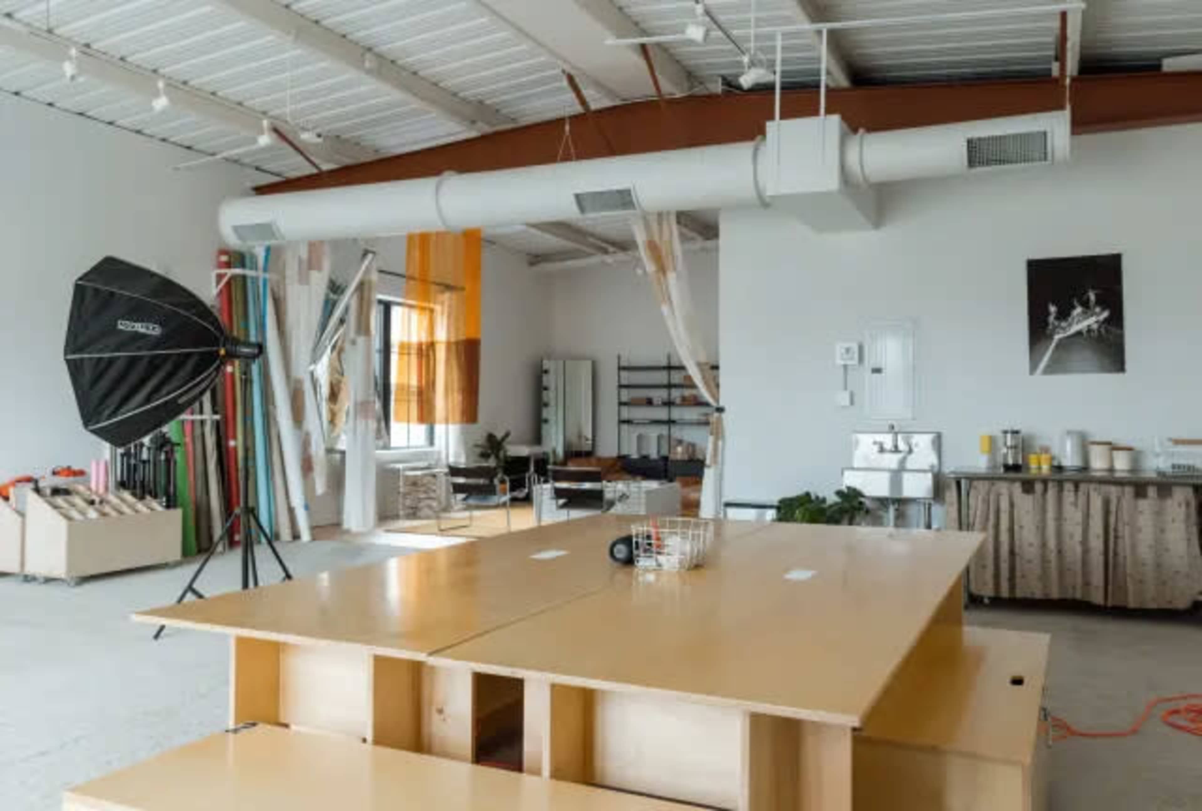 The image shows a spacious studio equipped with a large wooden table, photography equipment, and various shelving units against a bright, open backdrop.