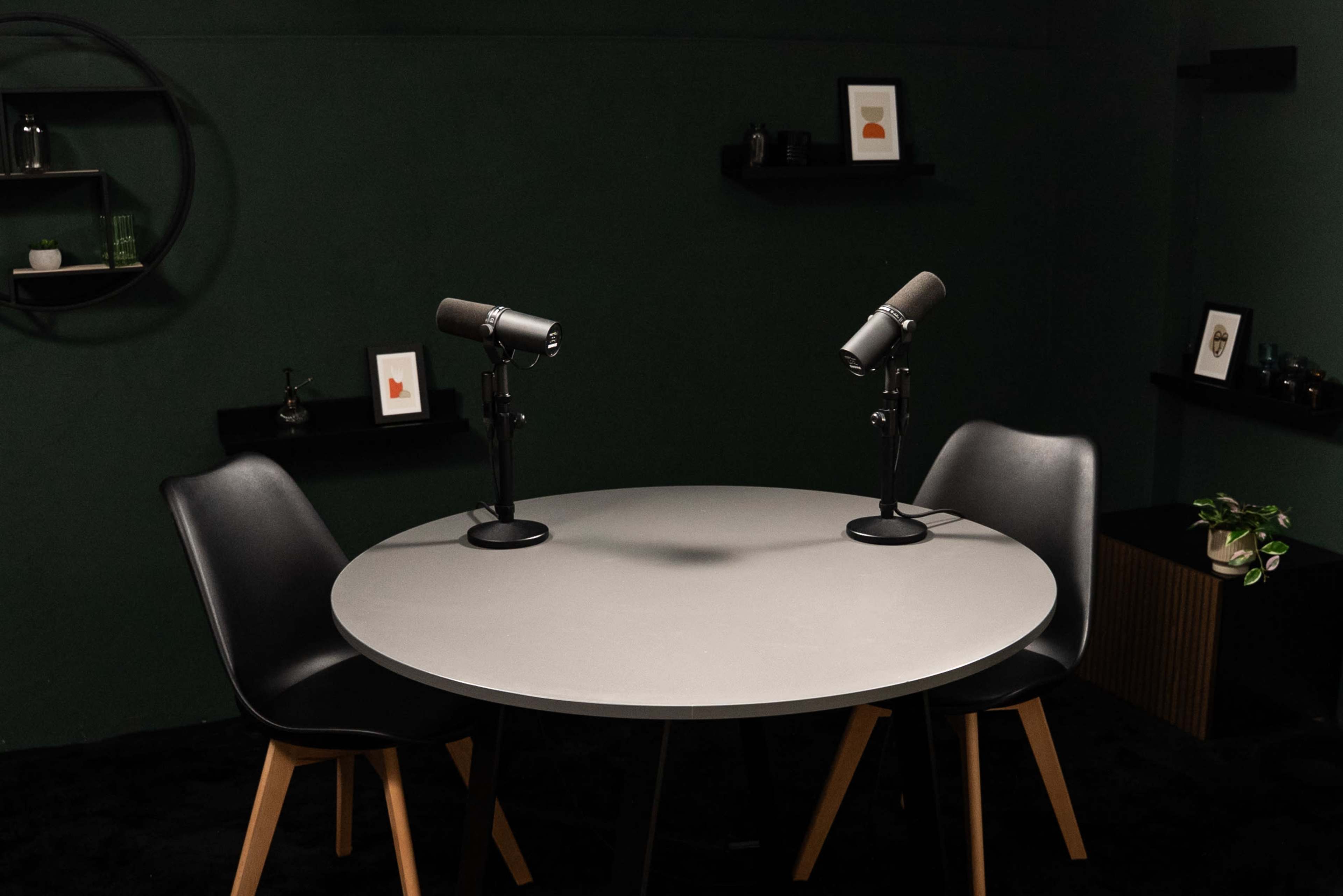 A round table with two microphones is set between two black chairs in a dark green room with minimal decorations on the walls.