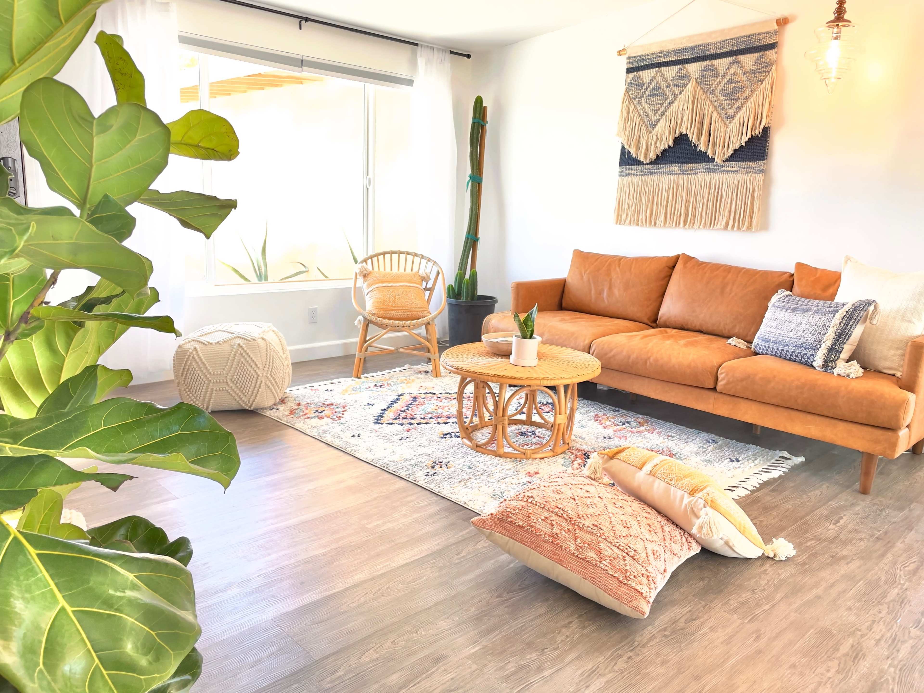 The image shows a bright living room featuring a brown leather sofa, a round wooden coffee table, woven decorations on the wall, and a patterned area rug.