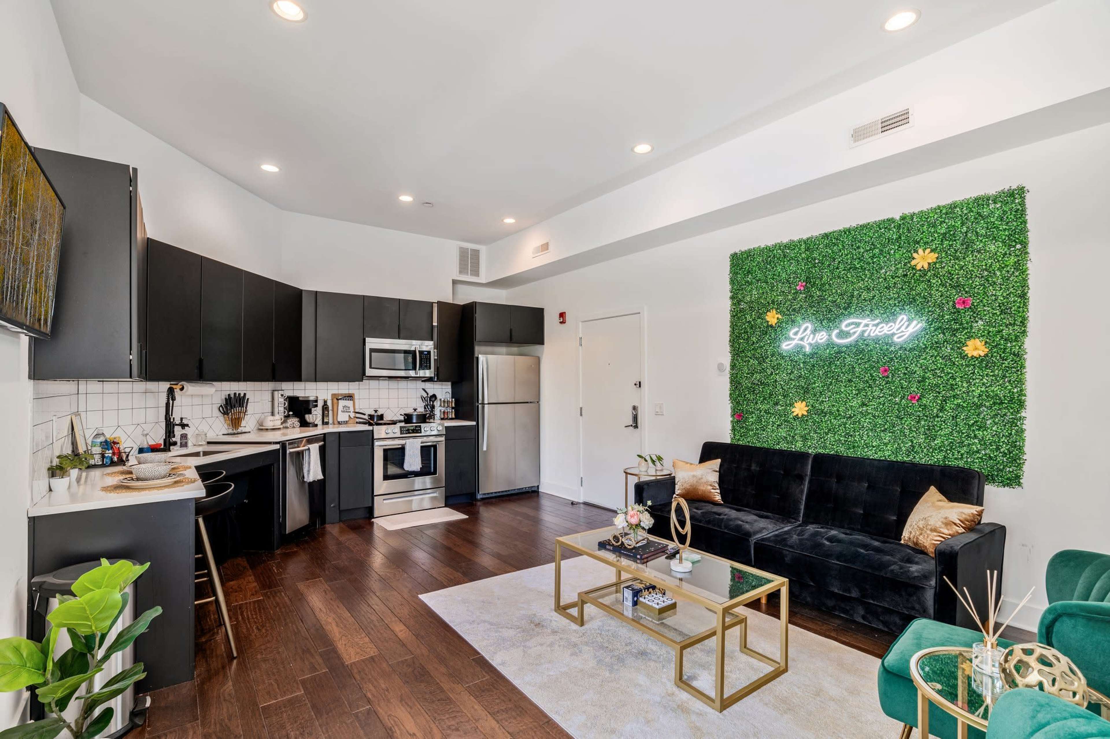 A modern kitchen and living area featuring dark cabinetry, stainless steel appliances, and a green wall with a neon sign that reads "Be Free."