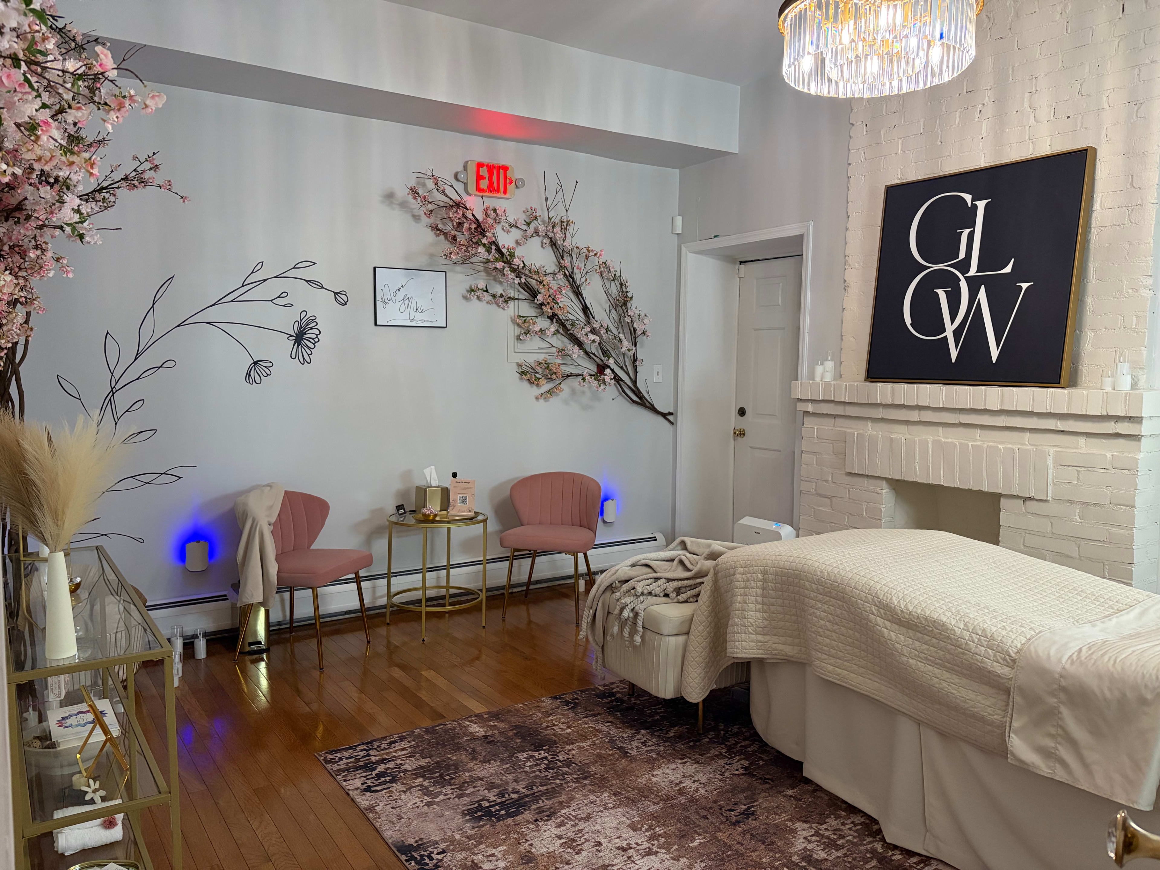 The image shows a cozy spa room featuring a treatment table, decorative pink chairs, floral wall art, and soft lighting, with a chandelier overhead.