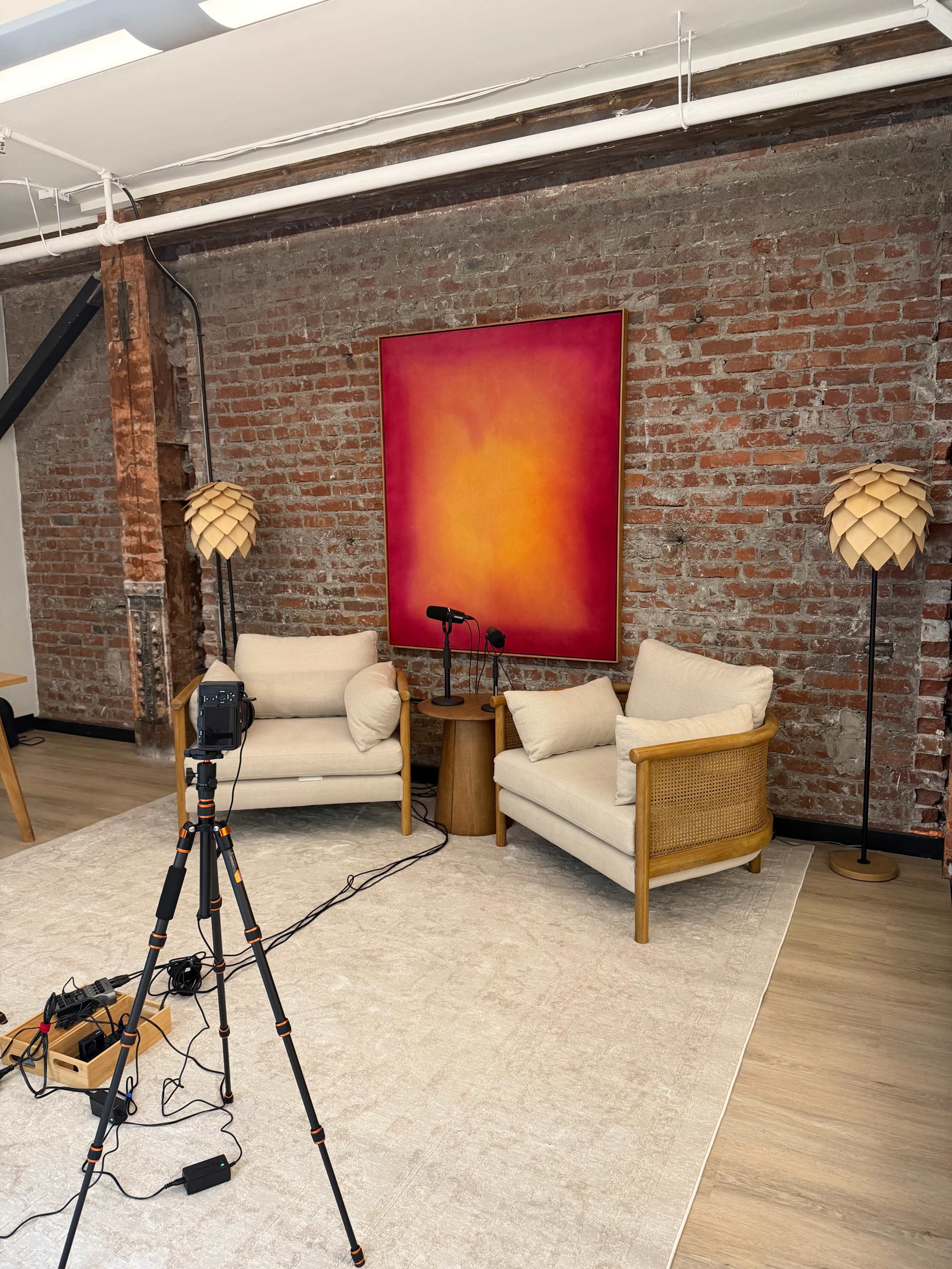 The image shows a well-lit studio space with two beige armchairs facing a small wooden table, a large pink and orange abstract painting on the brick wall behind, and a microphone setup in front.