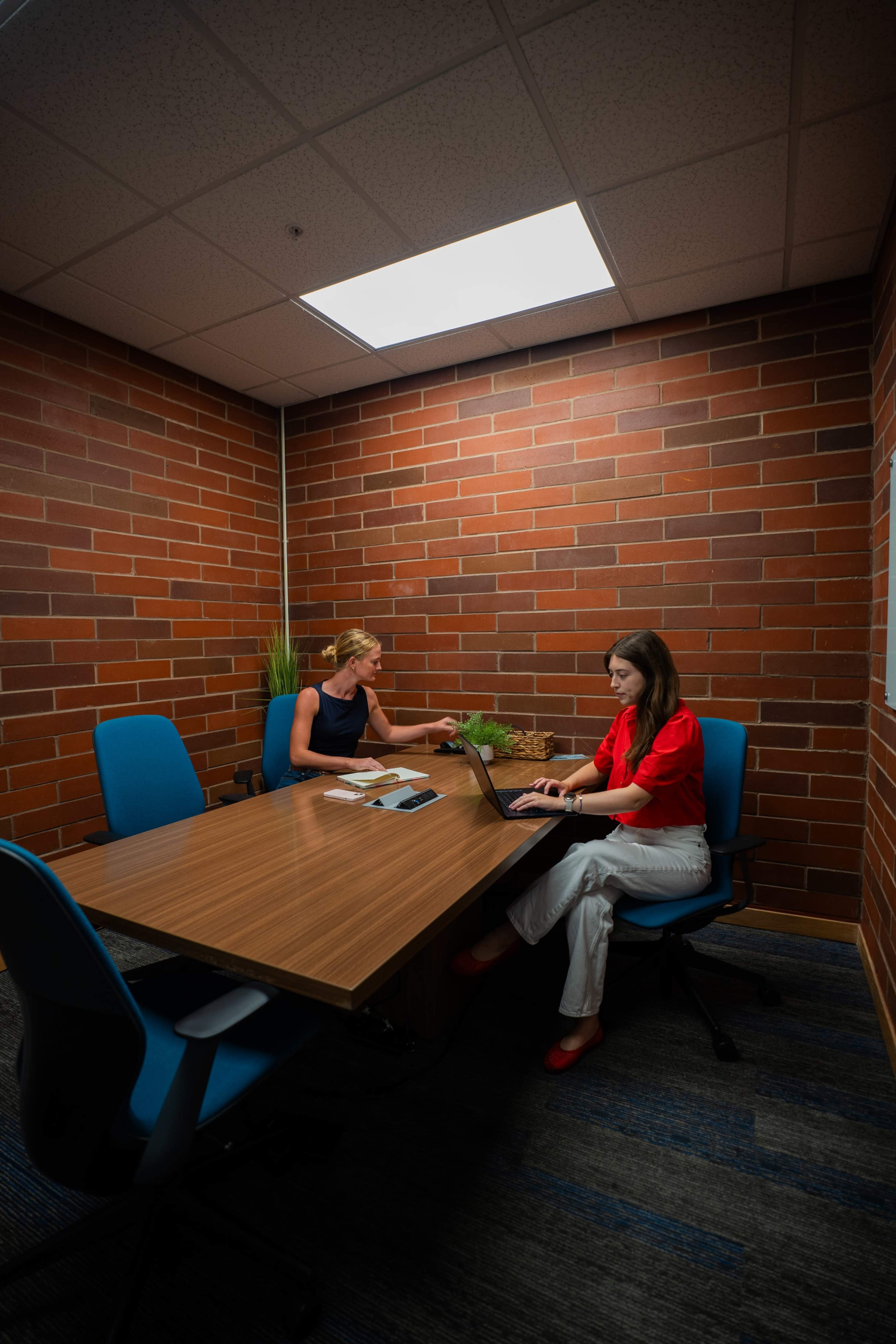 Two women are seated at a wooden table in a small conference room with brick walls, one working on a laptop and the other engaged in conversation.
