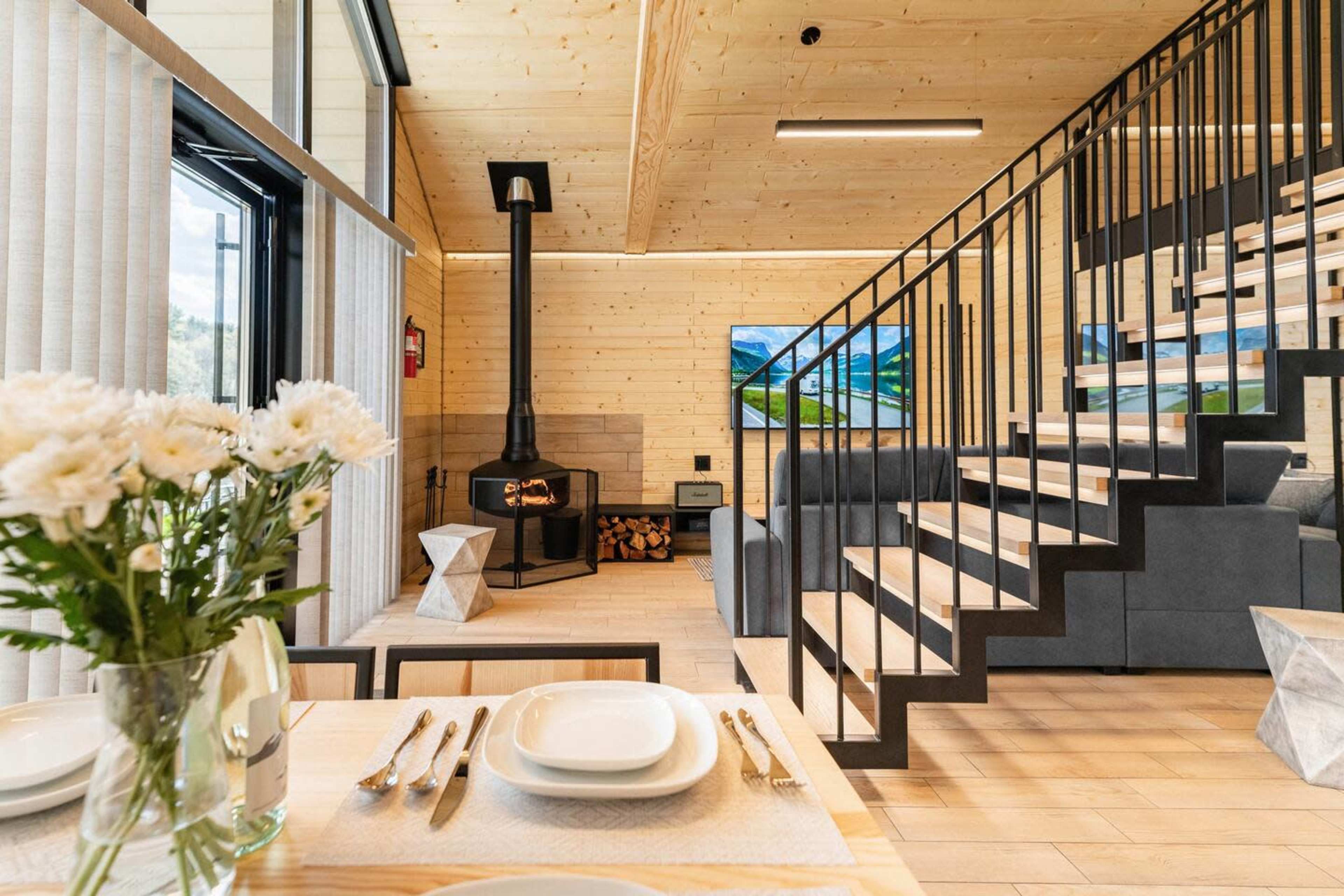A modern, wooden interior living space featuring a staircase, a fireplace, a dining table with flowers, and large windows.
