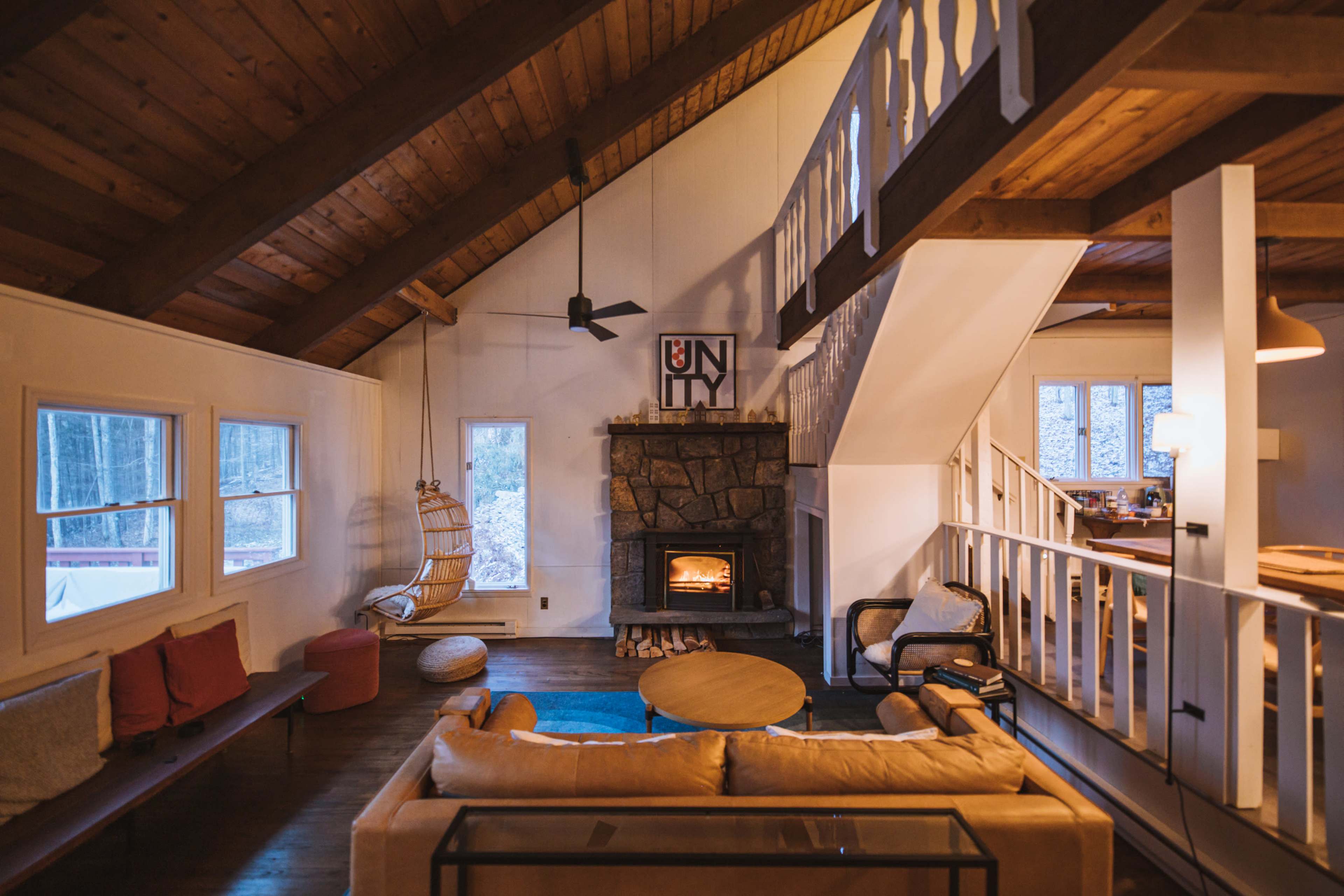 A cozy living room in a cabin with a stone fireplace, wooden beams, and a large window overlooking a wooded area.