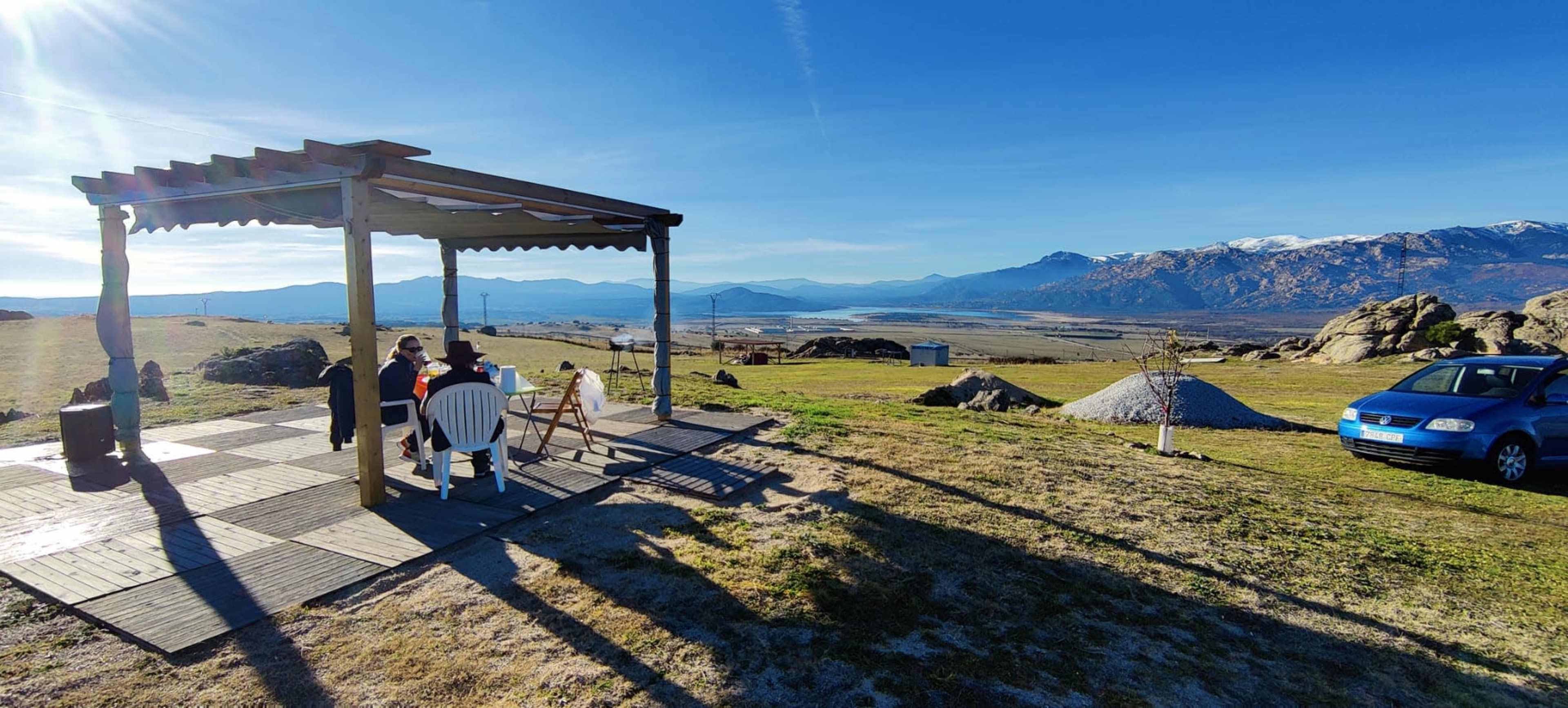 A person sits at a table under a wooden pergola, with a dog nearby and a car parked in the grass, overlooking a mountainous landscape in the distance.