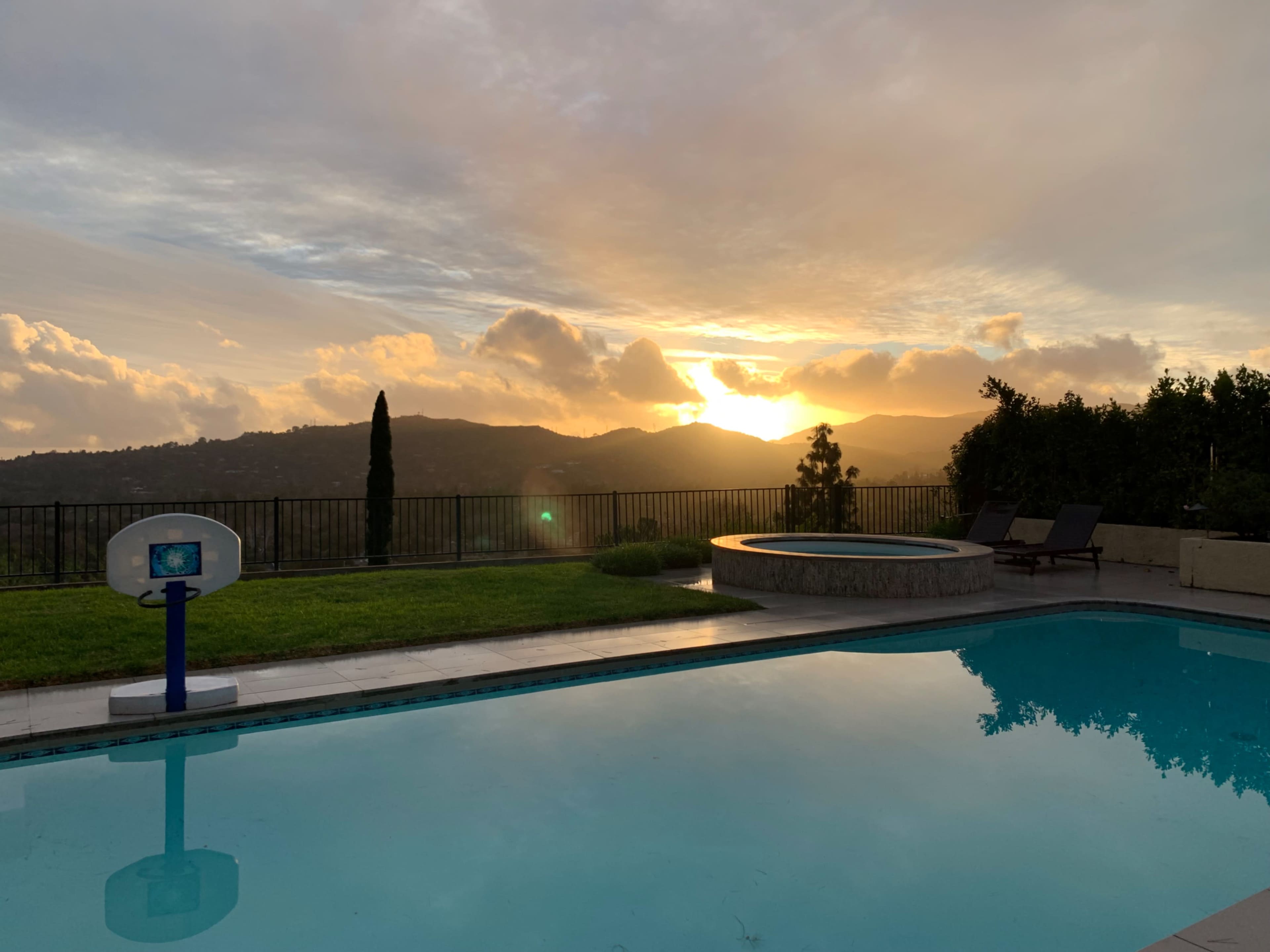 A swimming pool with a basketball hoop and a spa nearby, overlooking a scenic landscape at sunset.