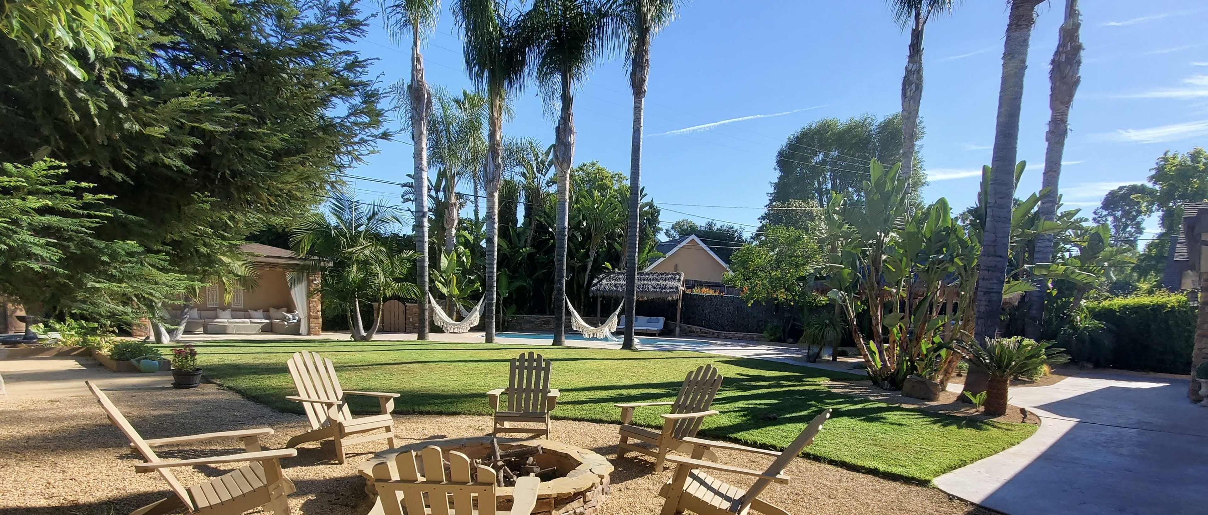 A backyard features a fire pit surrounded by several white adirondack chairs, with palm trees and a swimming pool in the background.