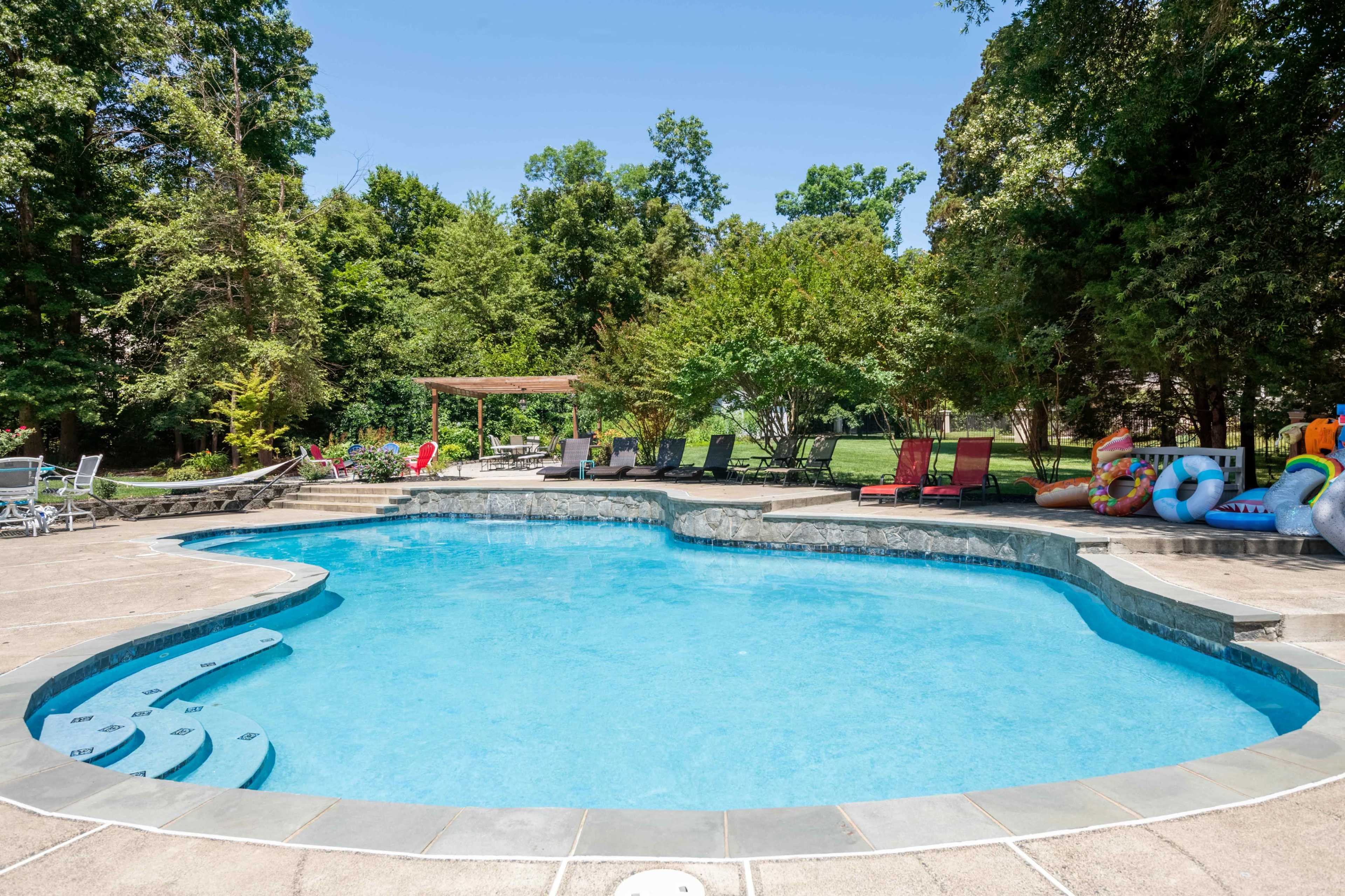 A backyard swimming pool surrounded by greenery and lounge chairs, with colorful pool floats arranged nearby.