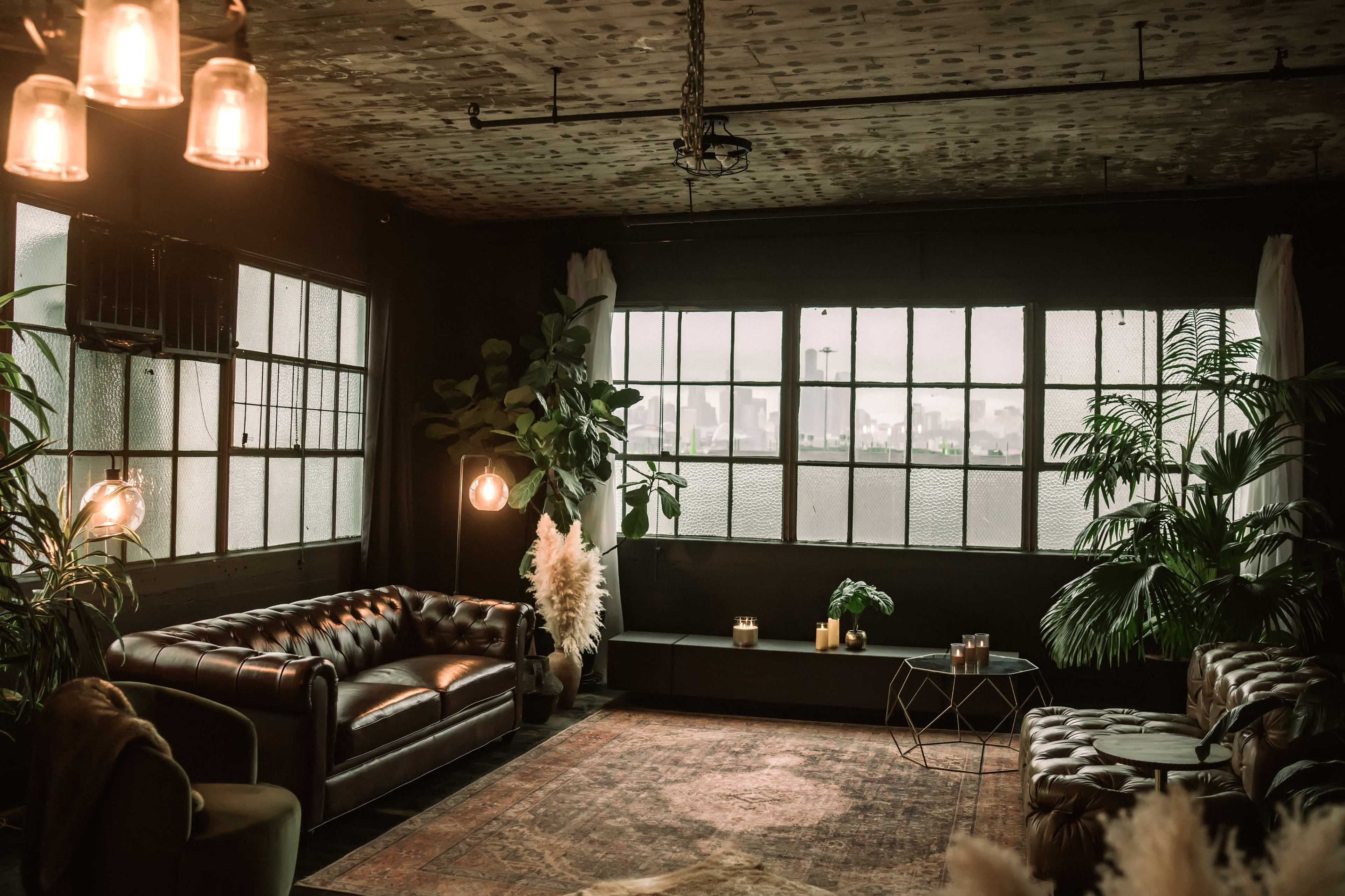 The image shows a stylish living space with a leather sofa, plants, and large windows allowing natural light to enter.