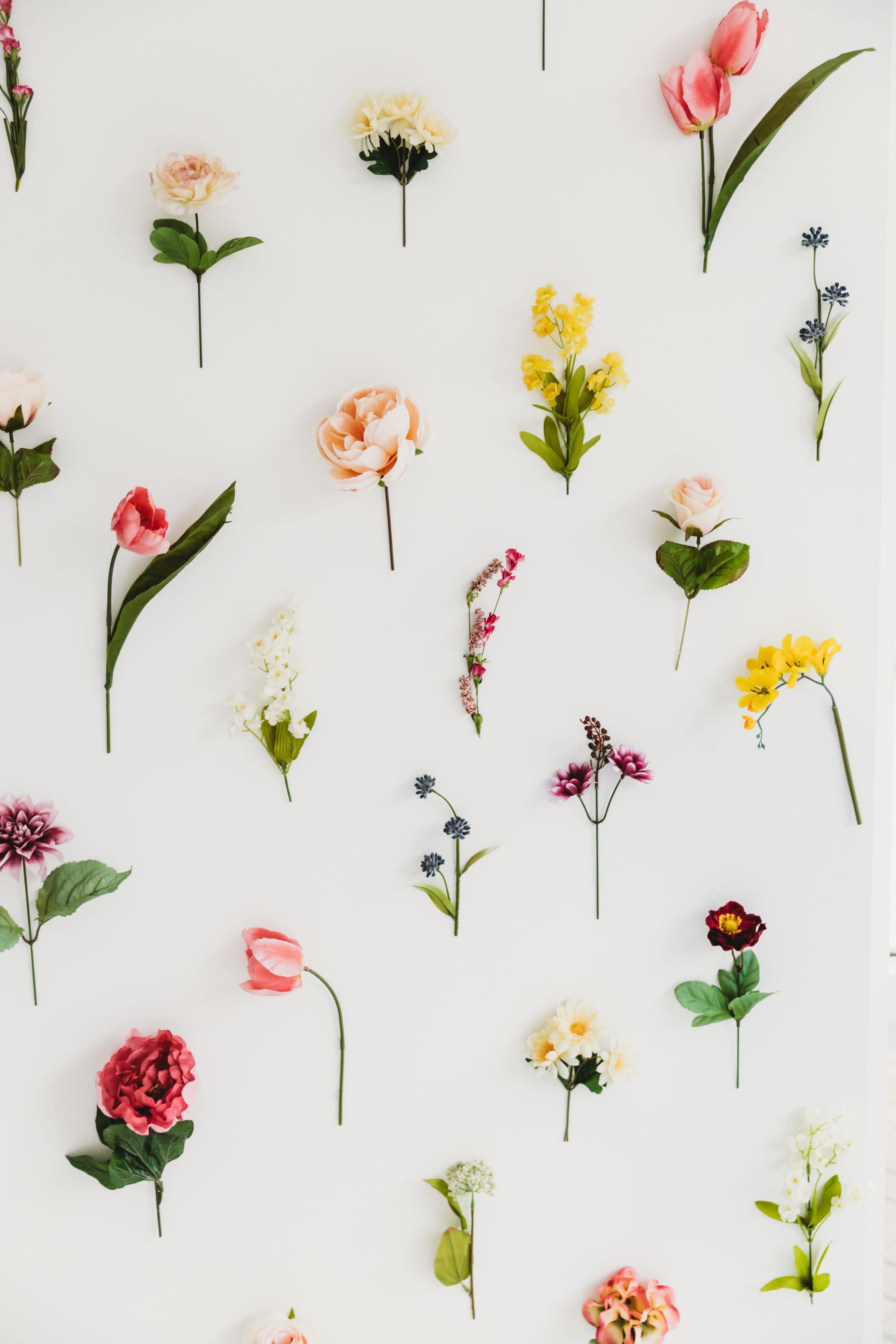 The image features a variety of artificial flowers arranged in a decorative pattern on a white wall.