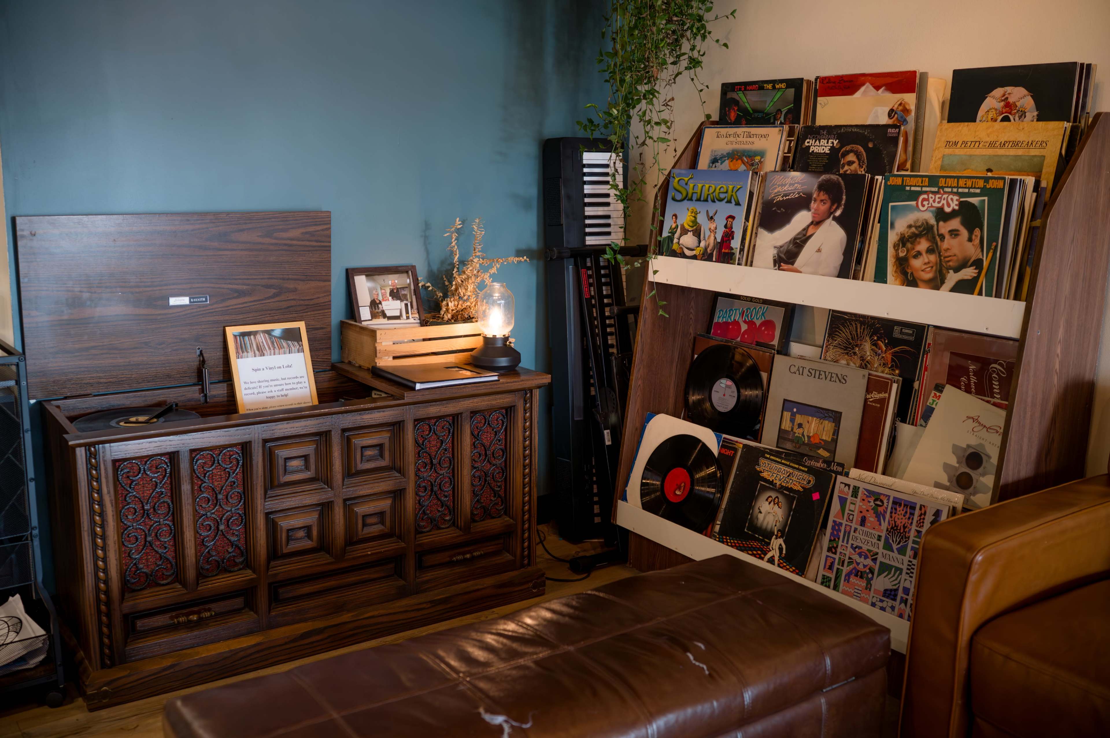 The image shows a vintage wooden record player next to a collection of vinyl records displayed on a white rack, with a small lamp and some decorative items on a nearby table.
