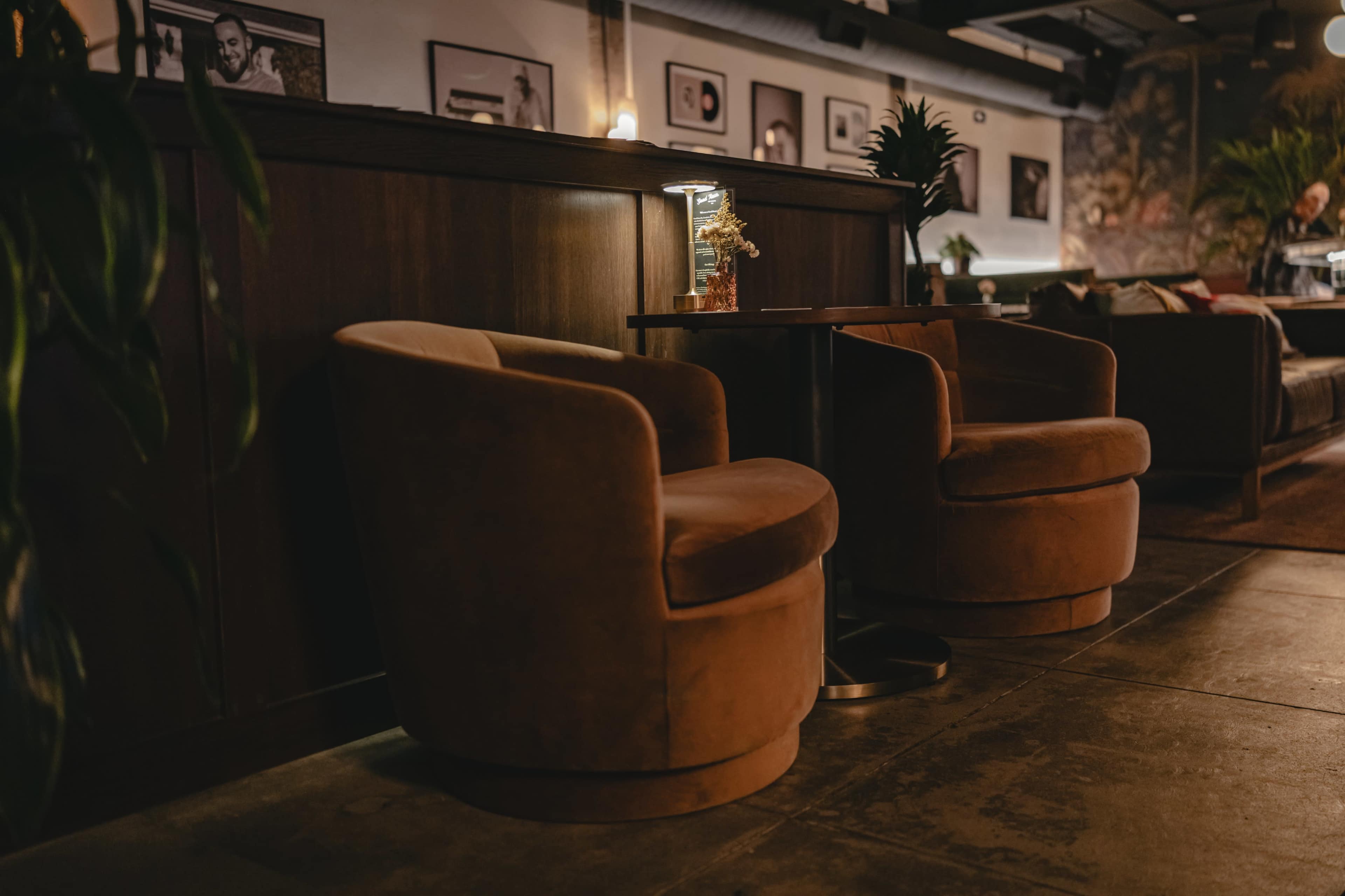 Two brown, upholstered armchairs are positioned near a small table in a cozy, dimly lit lounge adorned with framed pictures on the walls.