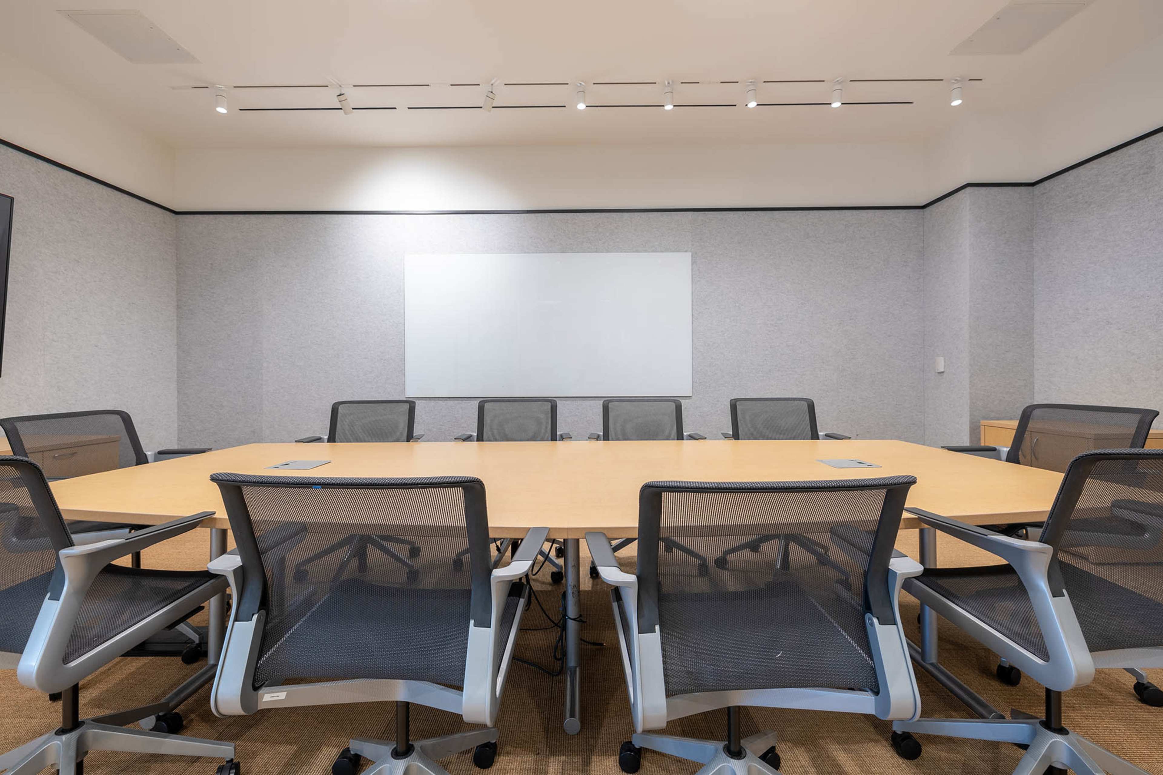 The image shows a conference room with a large circular wooden table surrounded by several ergonomic chairs and a whiteboard on the wall.