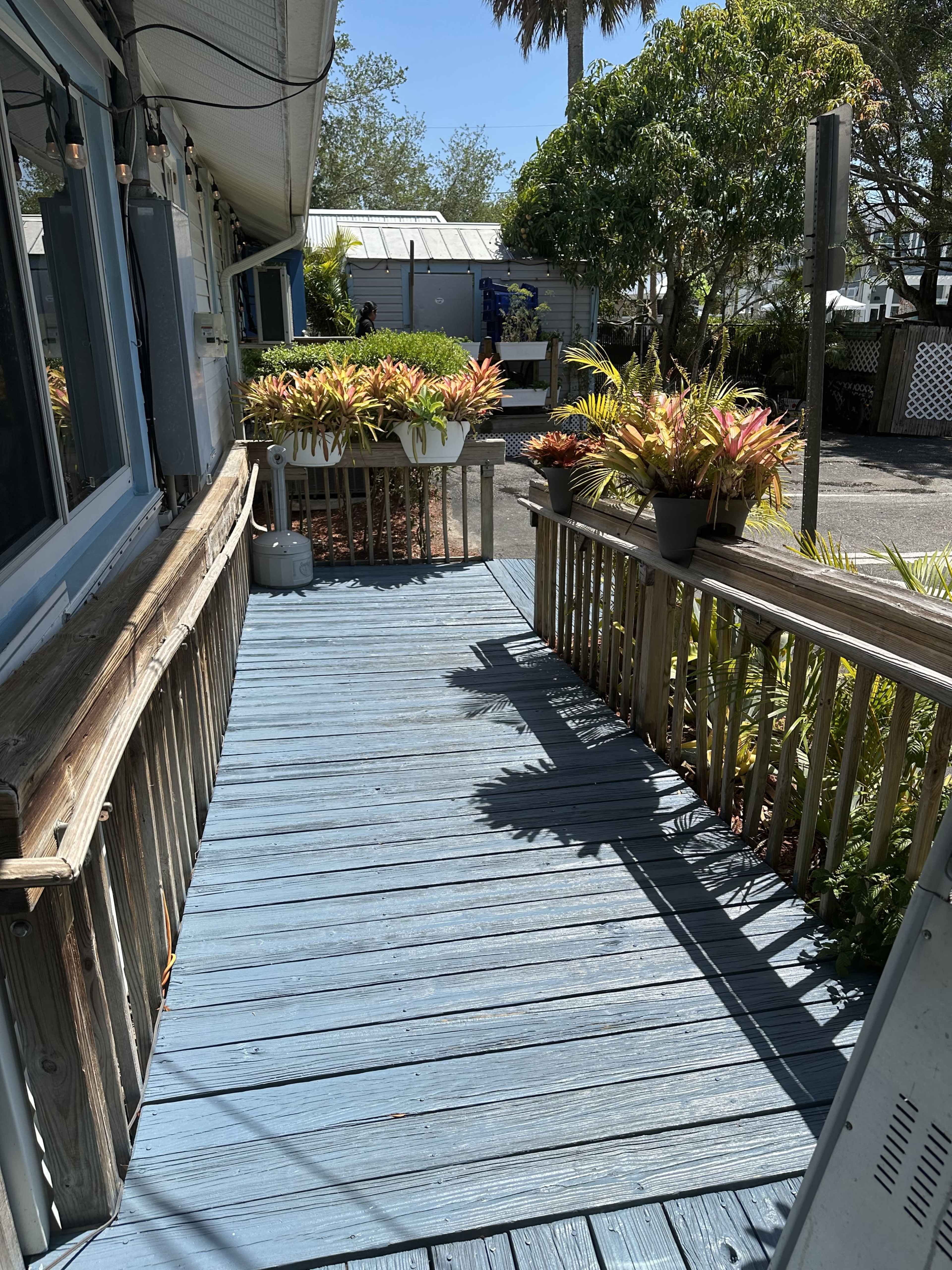 The image shows a blue-painted wooden deck lined with potted plants, leading to a building in a residential area.
