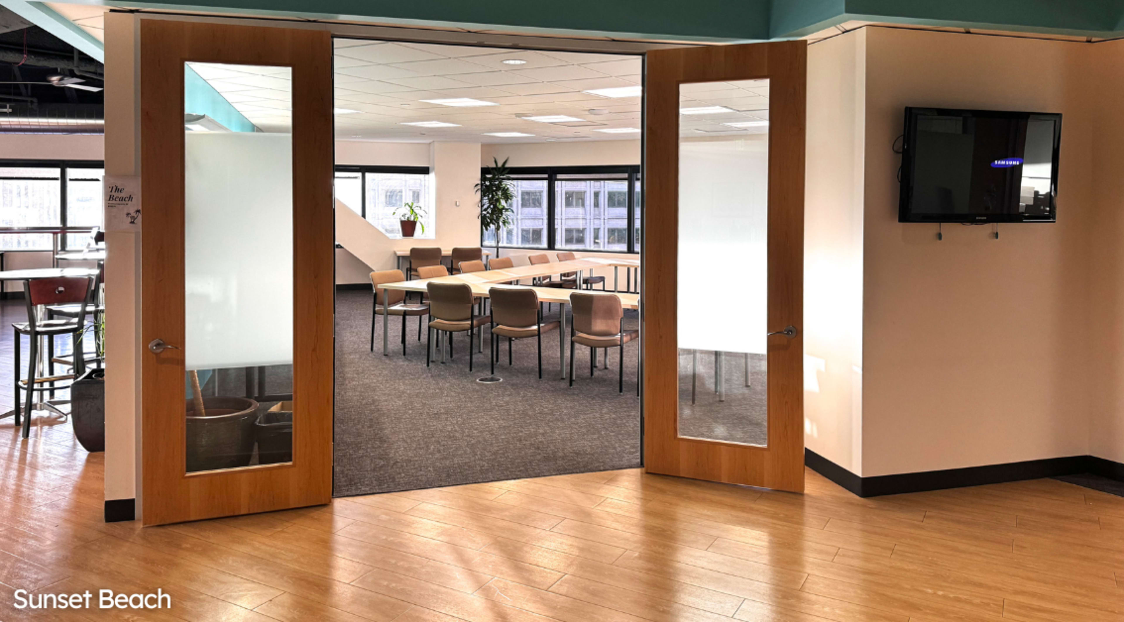 A large meeting room with a circular arrangement of chairs and a TV mounted on the wall, viewed through two glass doors.
