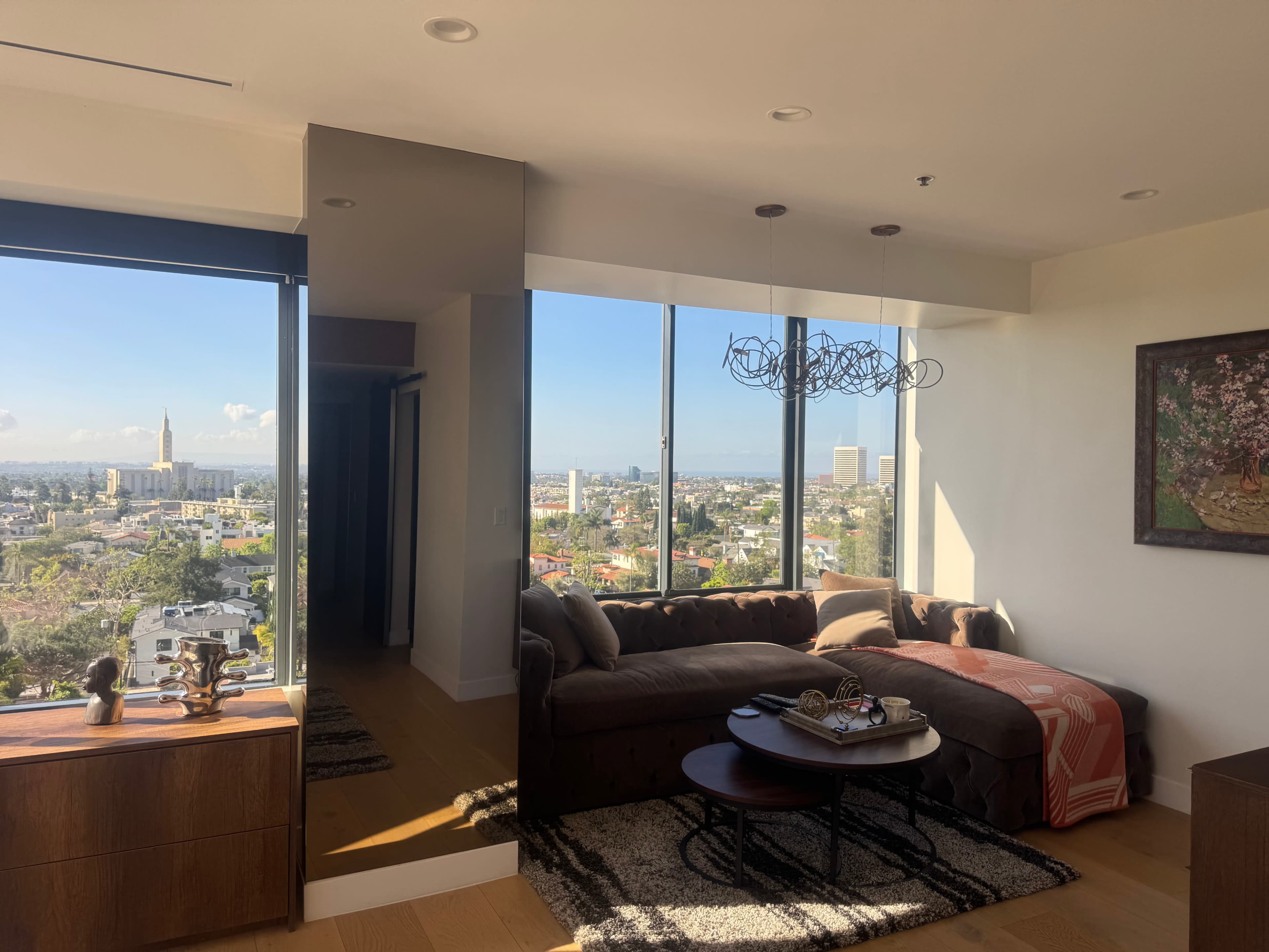 The image shows a modern living room with a large window offering a view of a cityscape, featuring a couch, a round coffee table, and a painting on the wall.