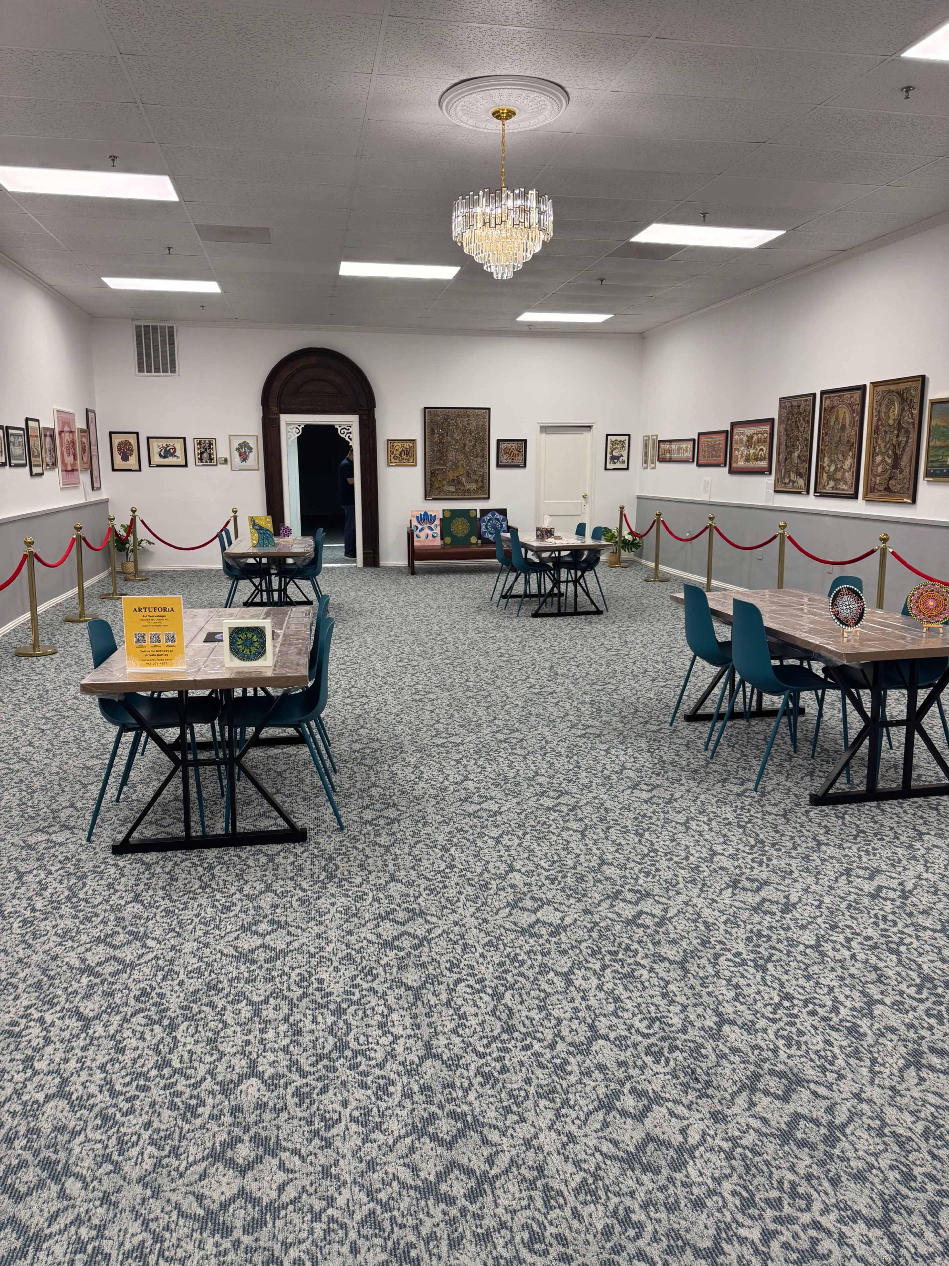 The image shows a spacious gallery room with multiple tables and chairs arranged throughout, featuring framed artwork on the walls and a chandelier hanging from the ceiling.