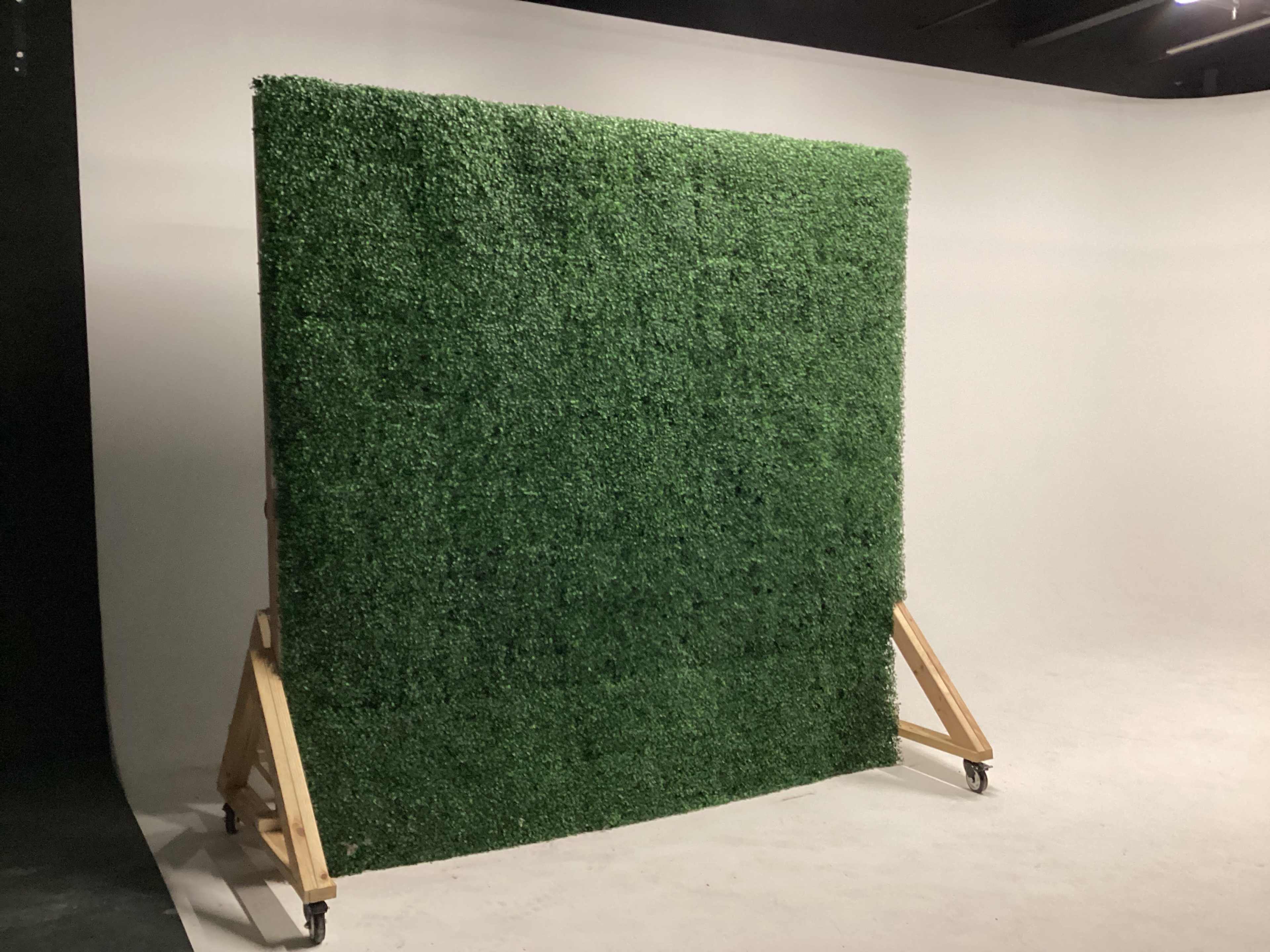 A large artificial grass wall stands on a wooden frame with wheels in a studio setting.